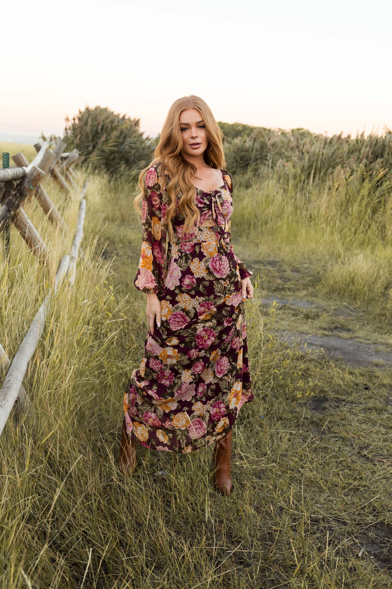 A floral dress flows gracefully in the golden evening light as a woman poses among tall grasses near a rustic fence.