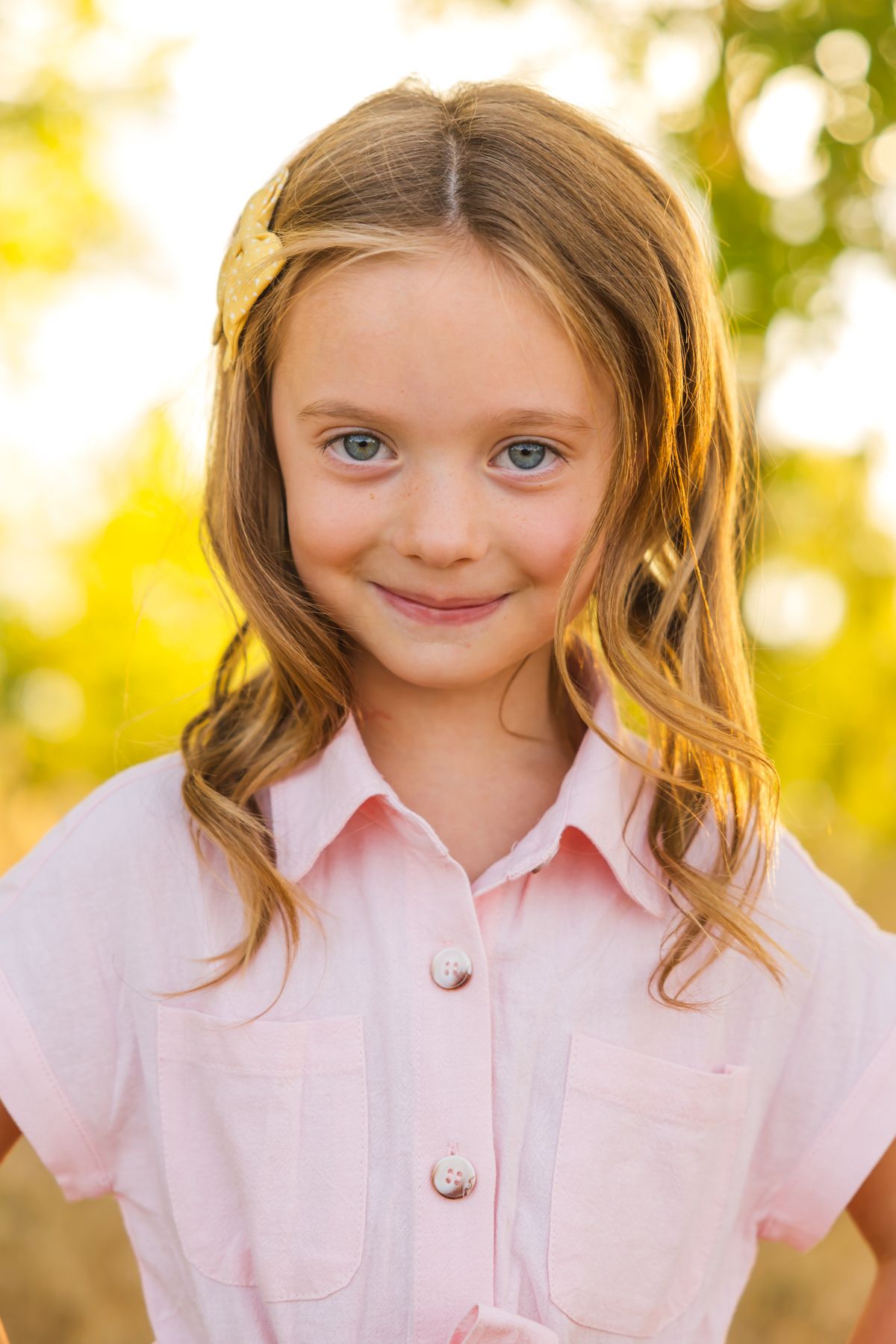 Portrait of a young person in a pink collared shirt with a yellow hair clip against a sunlit background.