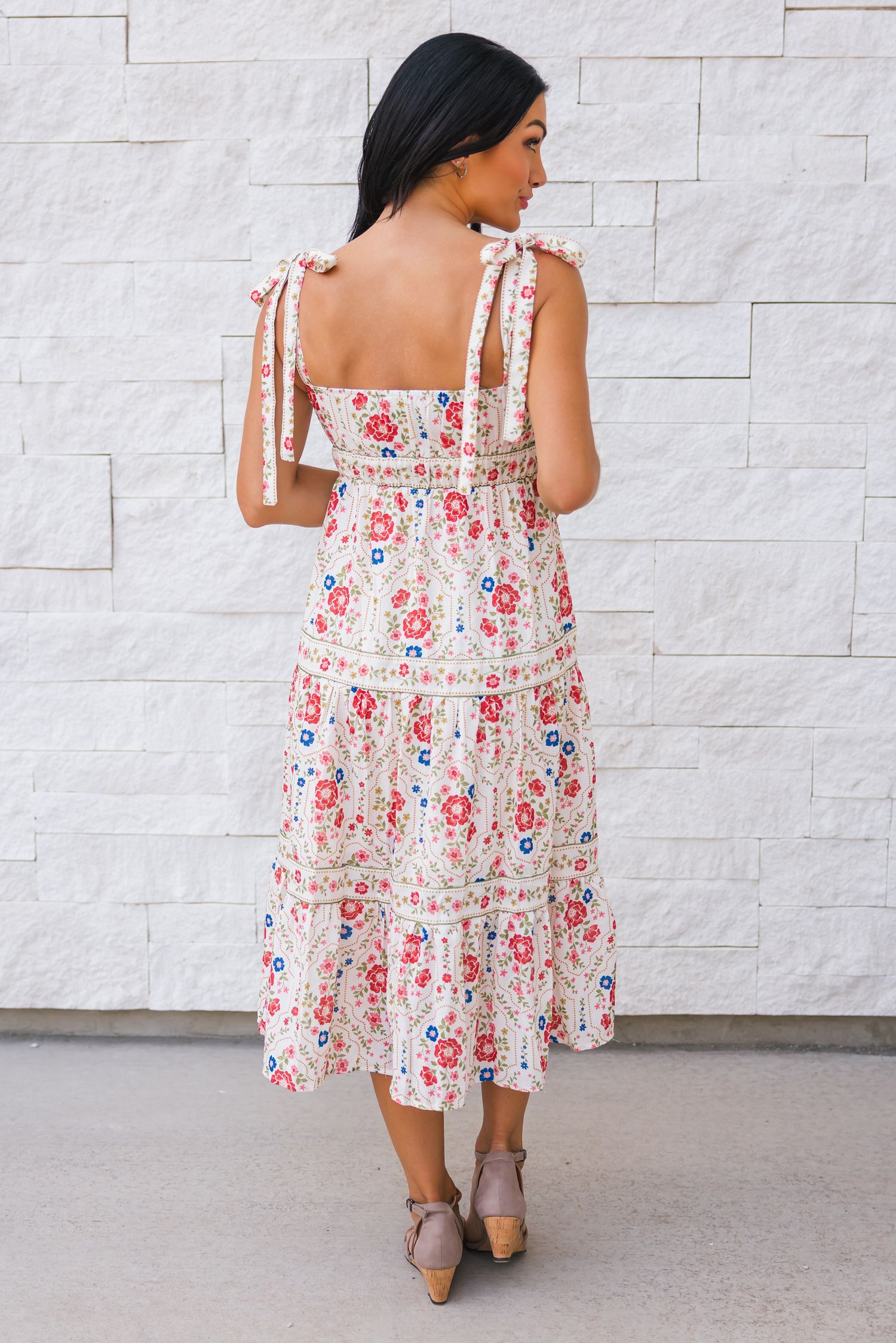 Woman wearing a floral print summer dress with shoulder ties poses against a white stone wall in three different angles.