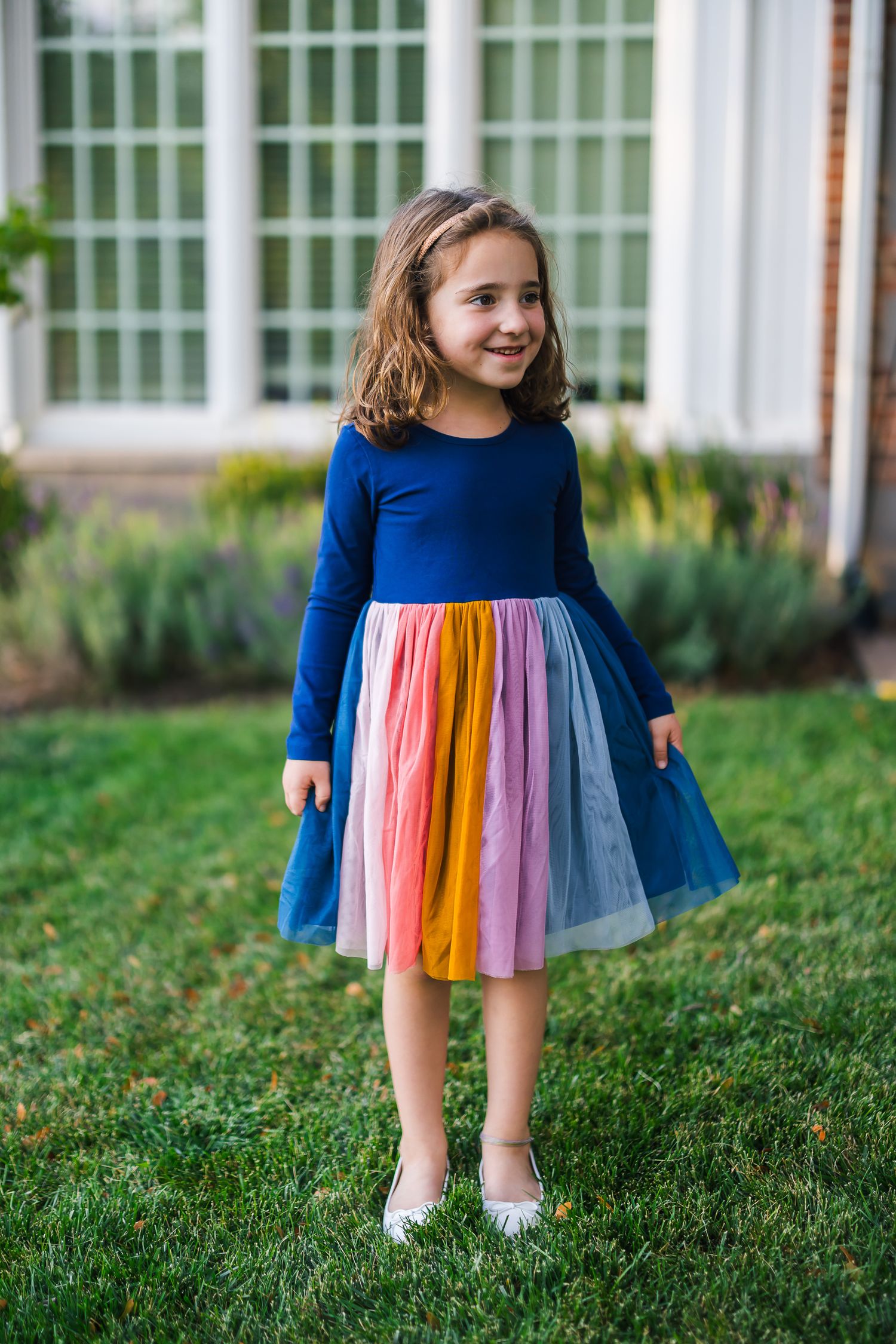 Young girl in a navy and rainbow dress poses against a white window backdrop.