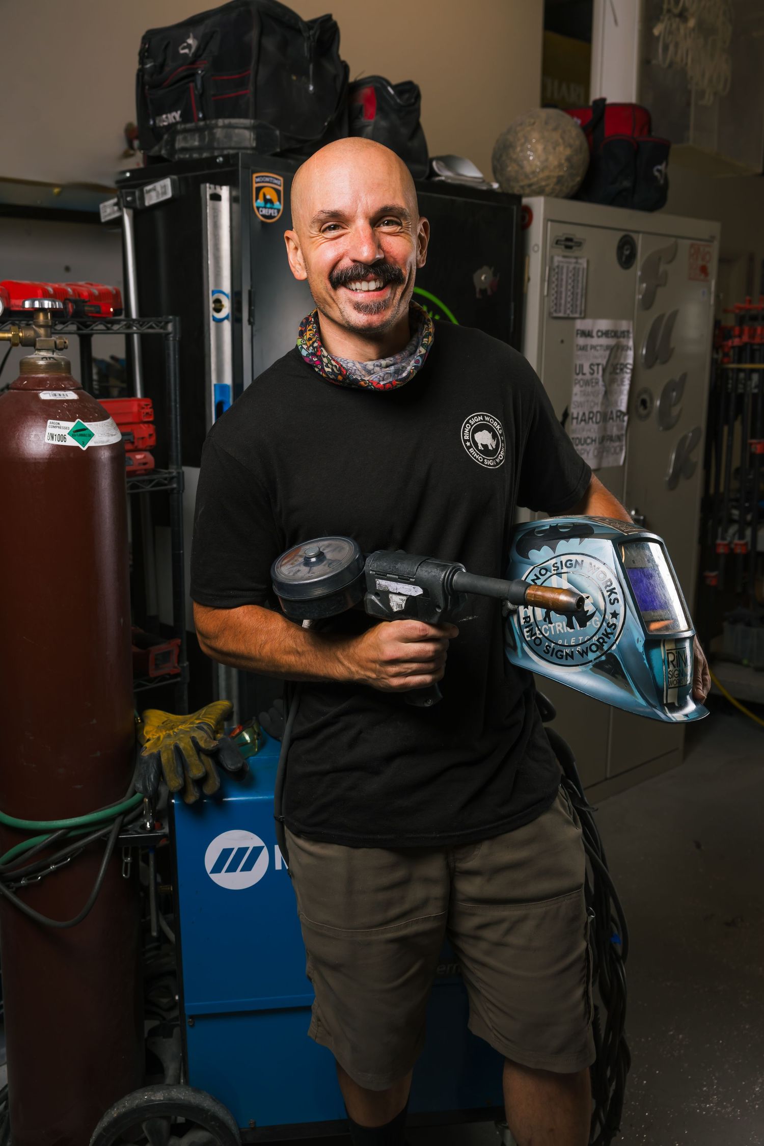Person in a black t-shirt holding auto repair tools smiles in a garage workshop with equipment in the background.