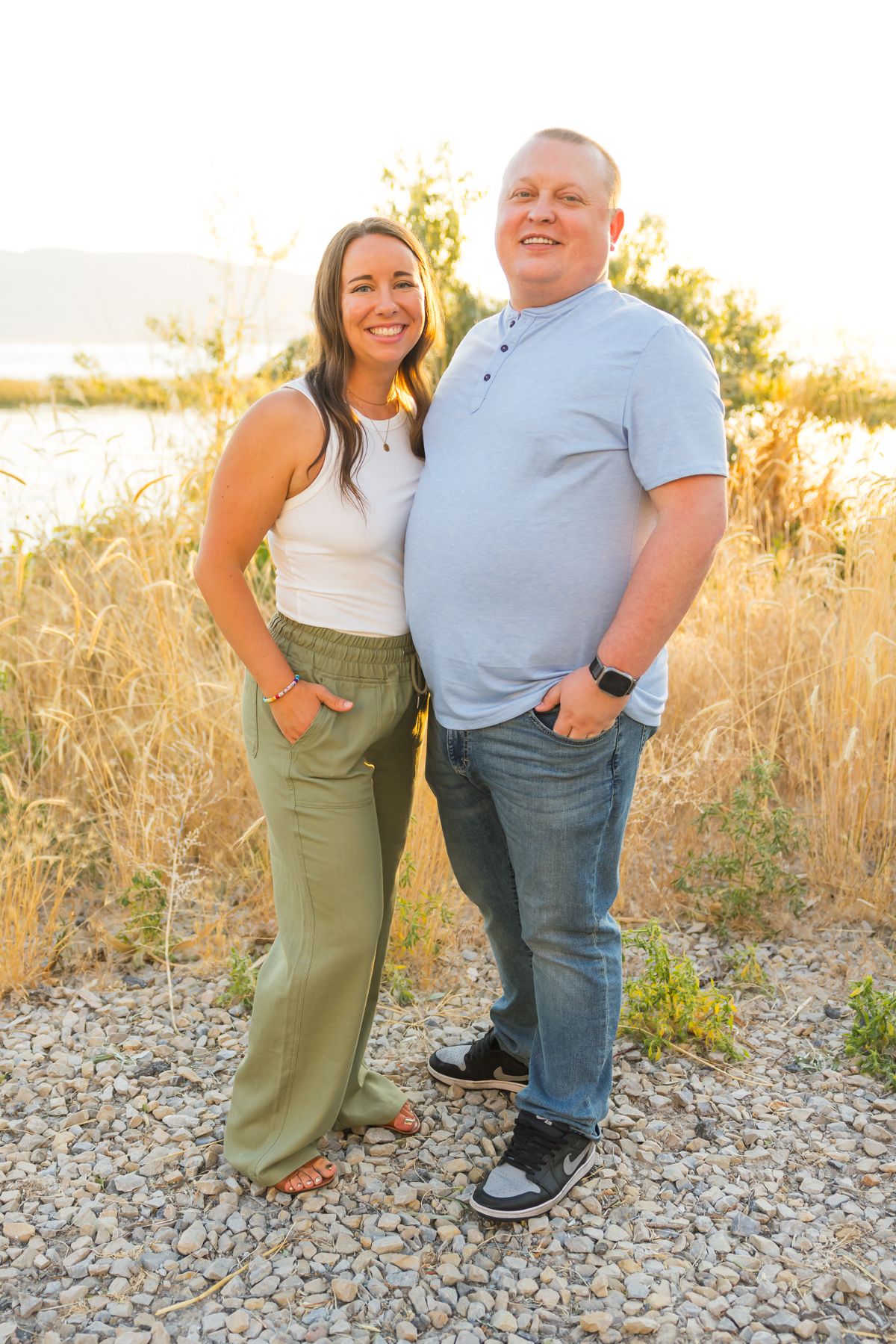 Couple in casual attire poses together on rocky path with water and sunset in background.