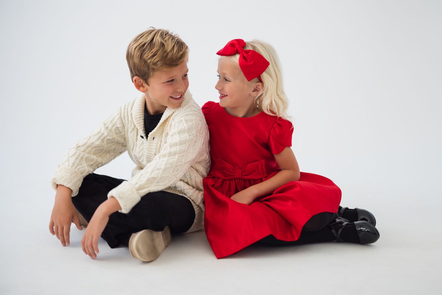 Two children in holiday attire, one in a cream cable knit sweater and one in a red dress with bow, sitting together.