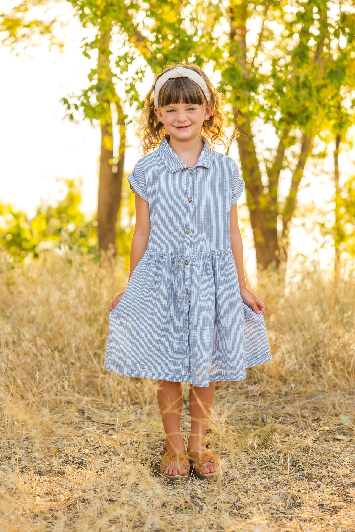 Child wearing blue dress twirls in sunlit field with golden light filtering through trees.