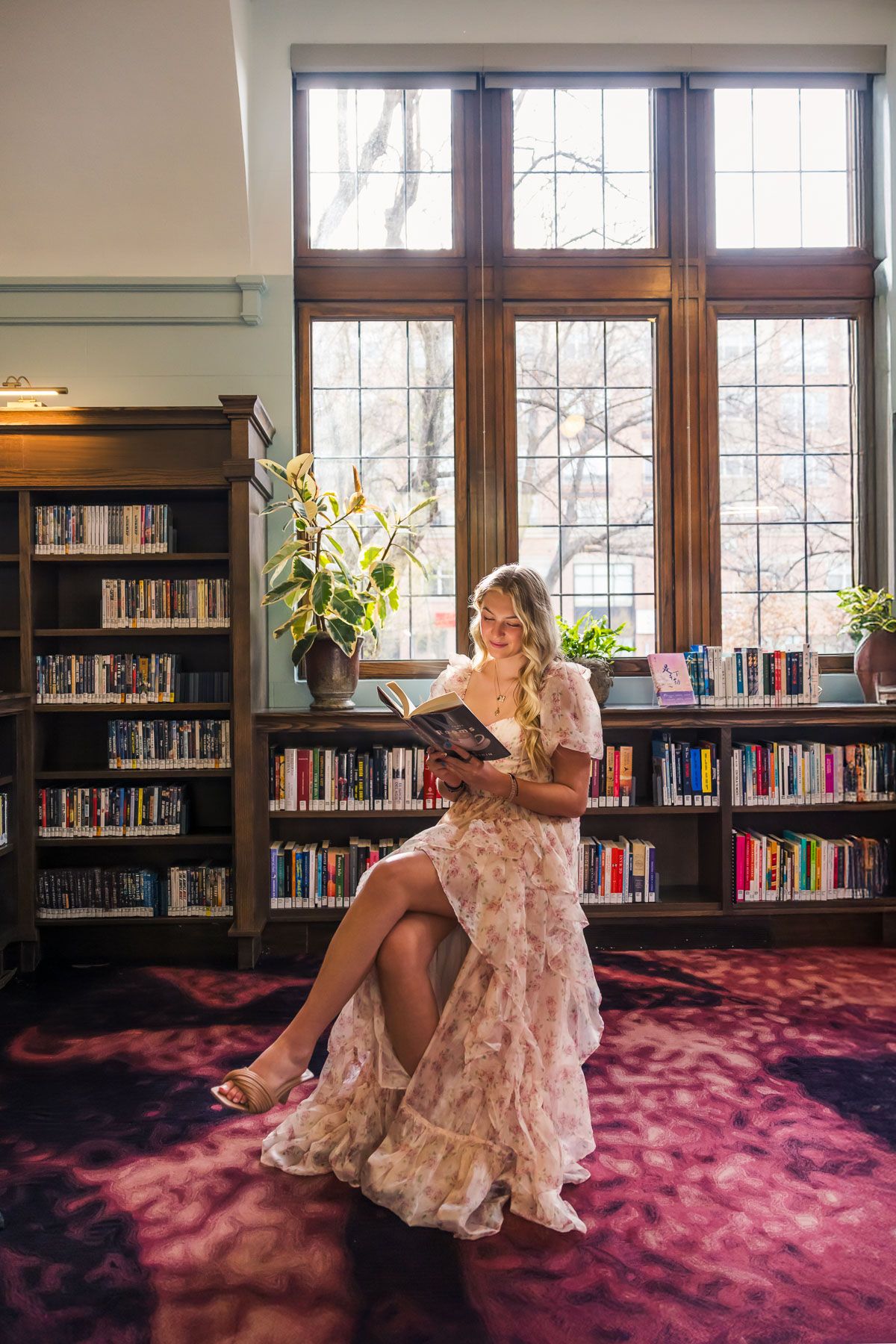Person in flowing dress reads book while sitting on pink carpet in library with wooden windows.