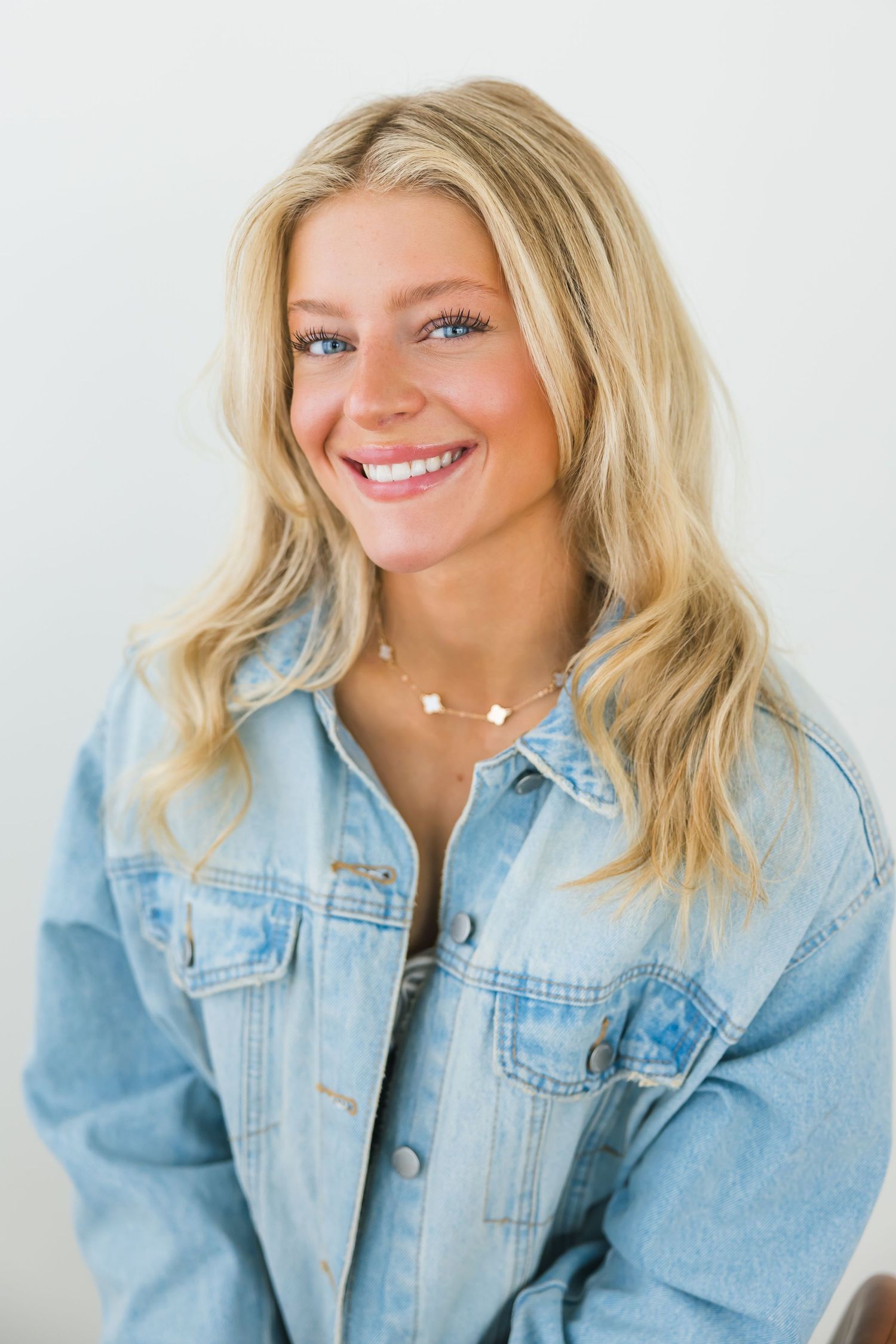 Smiling blonde woman in denim jacket wearing a delicate necklace against a light background.