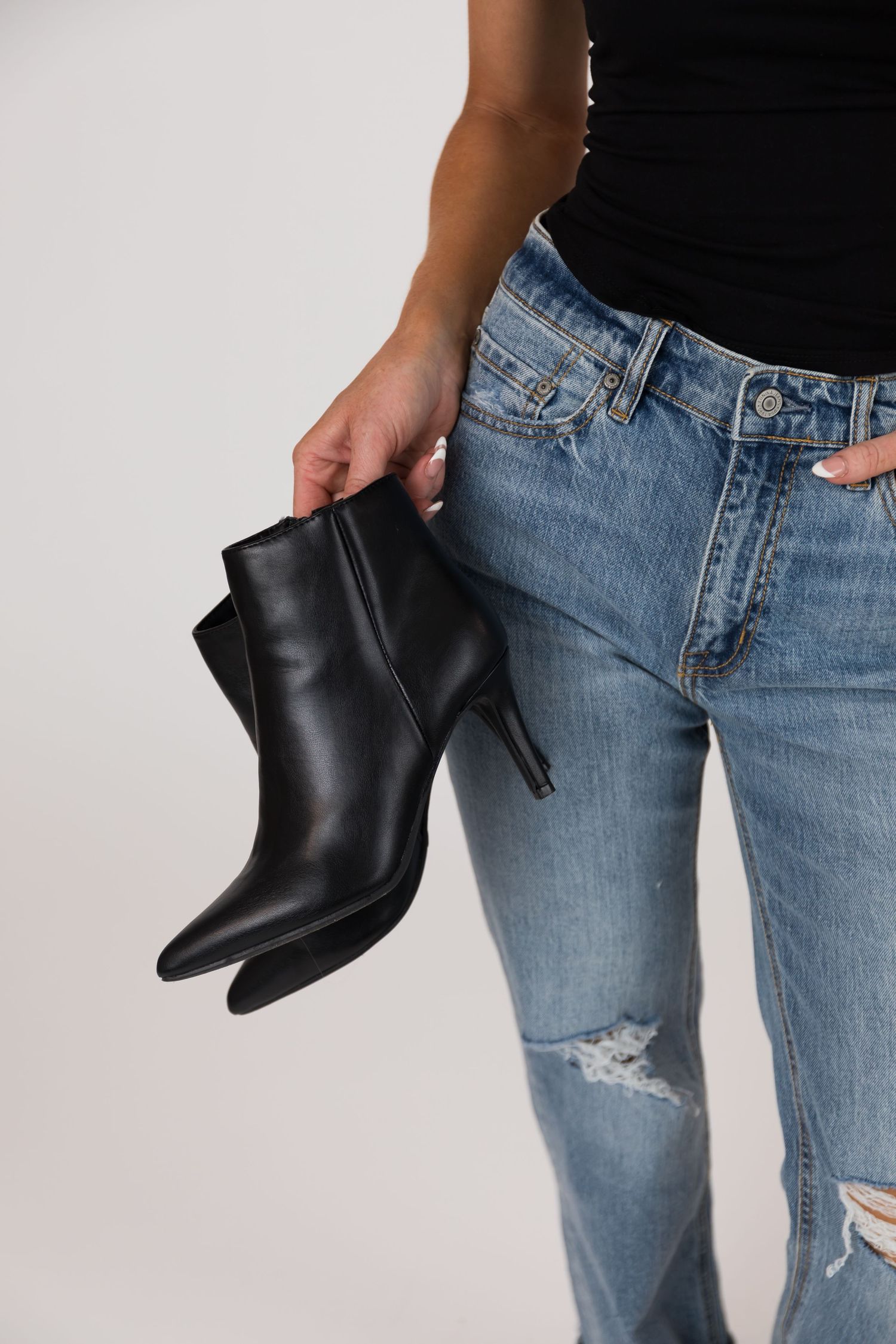 Woman in blue ripped jeans holding black leather ankle boots against white background.