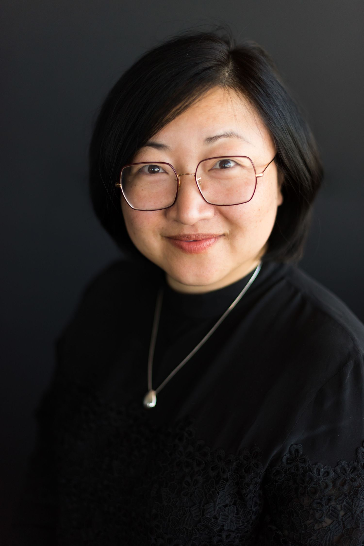 Professional headshot of a person wearing glasses and black attire with pendant necklace.