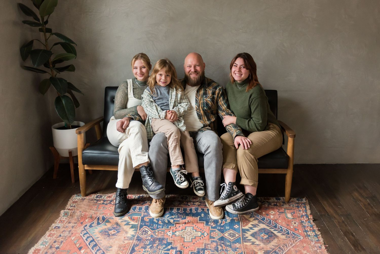 A group of four sits together on a blue couch over a patterned area rug against a gray wall with a potted plant.