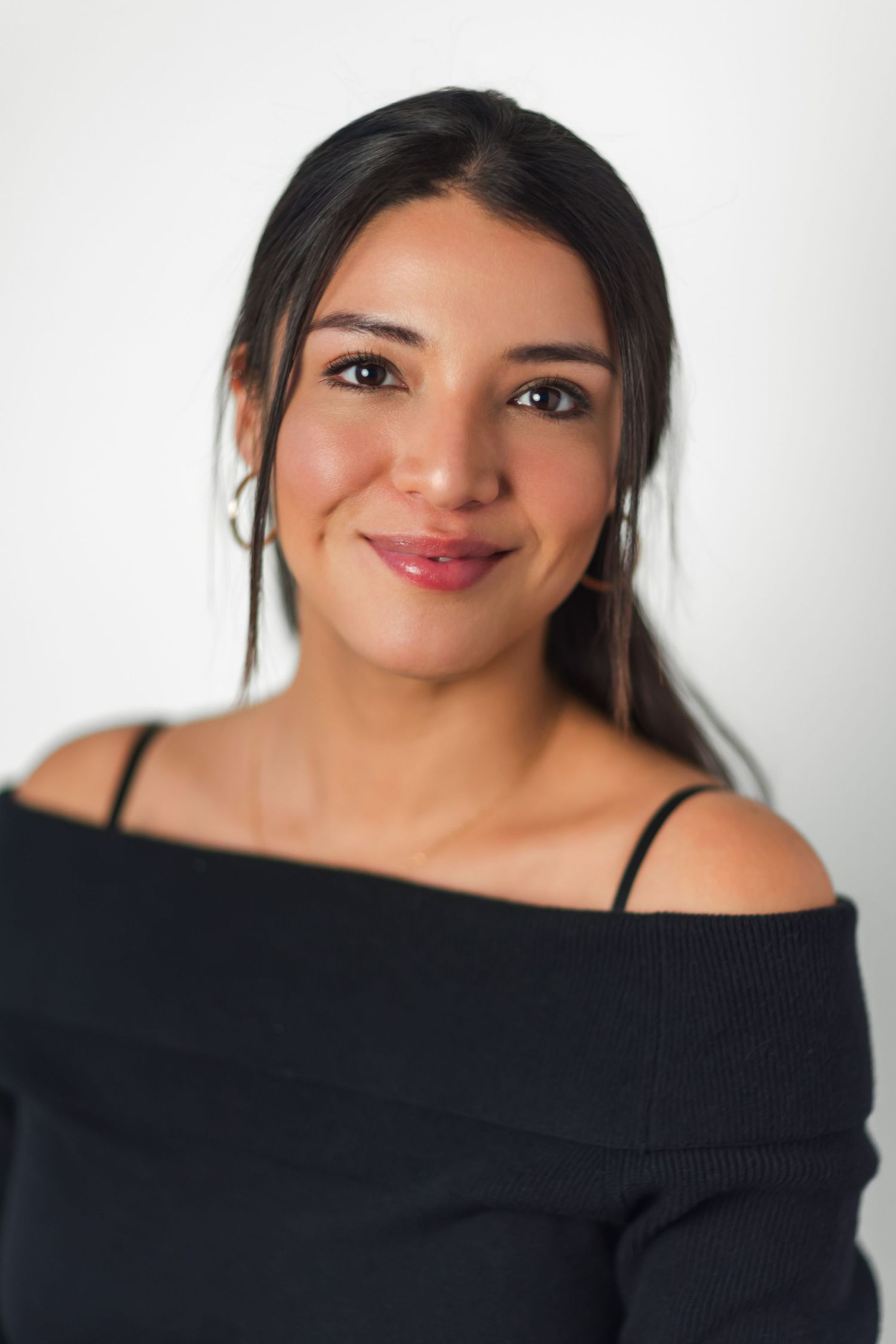 Portrait of someone with dark hair and a warm smile wearing an off-shoulder black top against a light background.