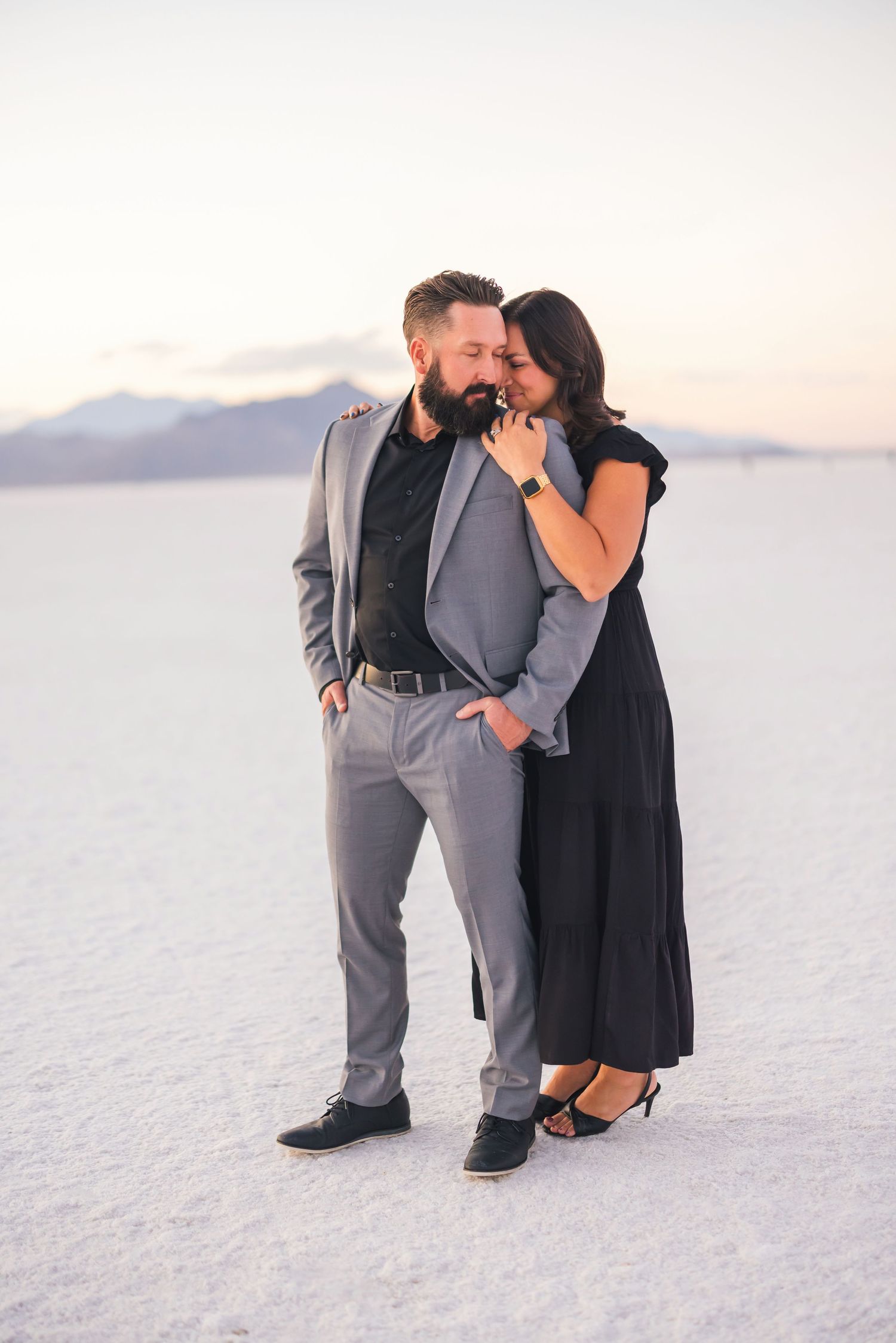Couple in formal attire poses playfully on white salt flats at sunset.
