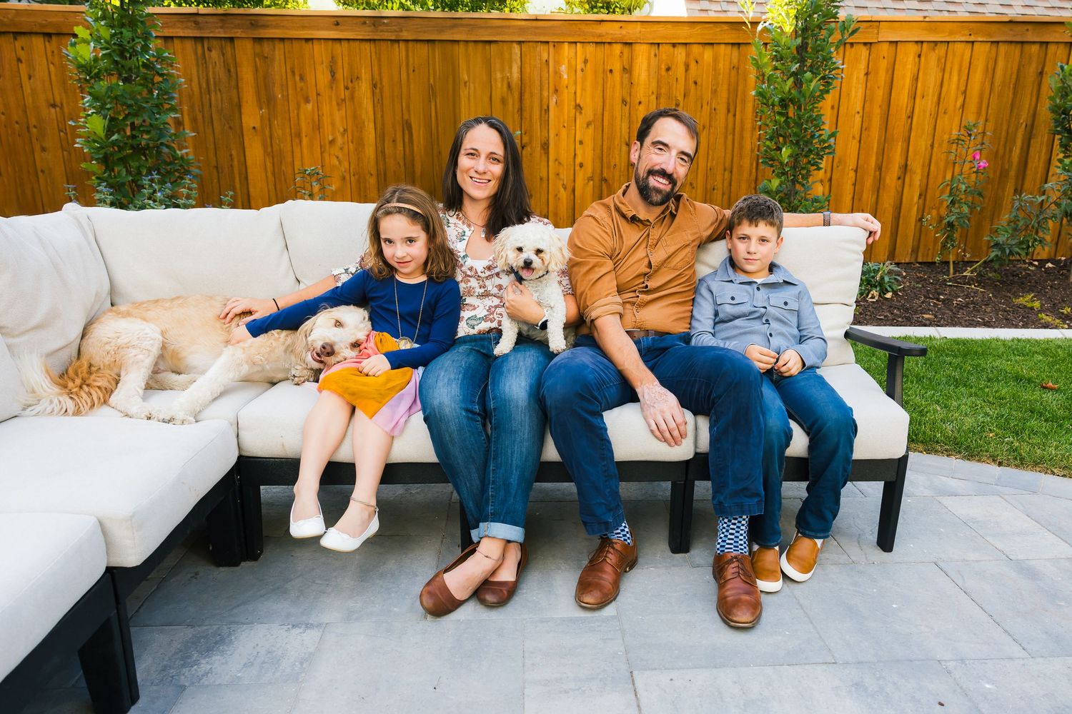 Family sitting together with dogs on sectional outdoor couch in backyard patio setting.
