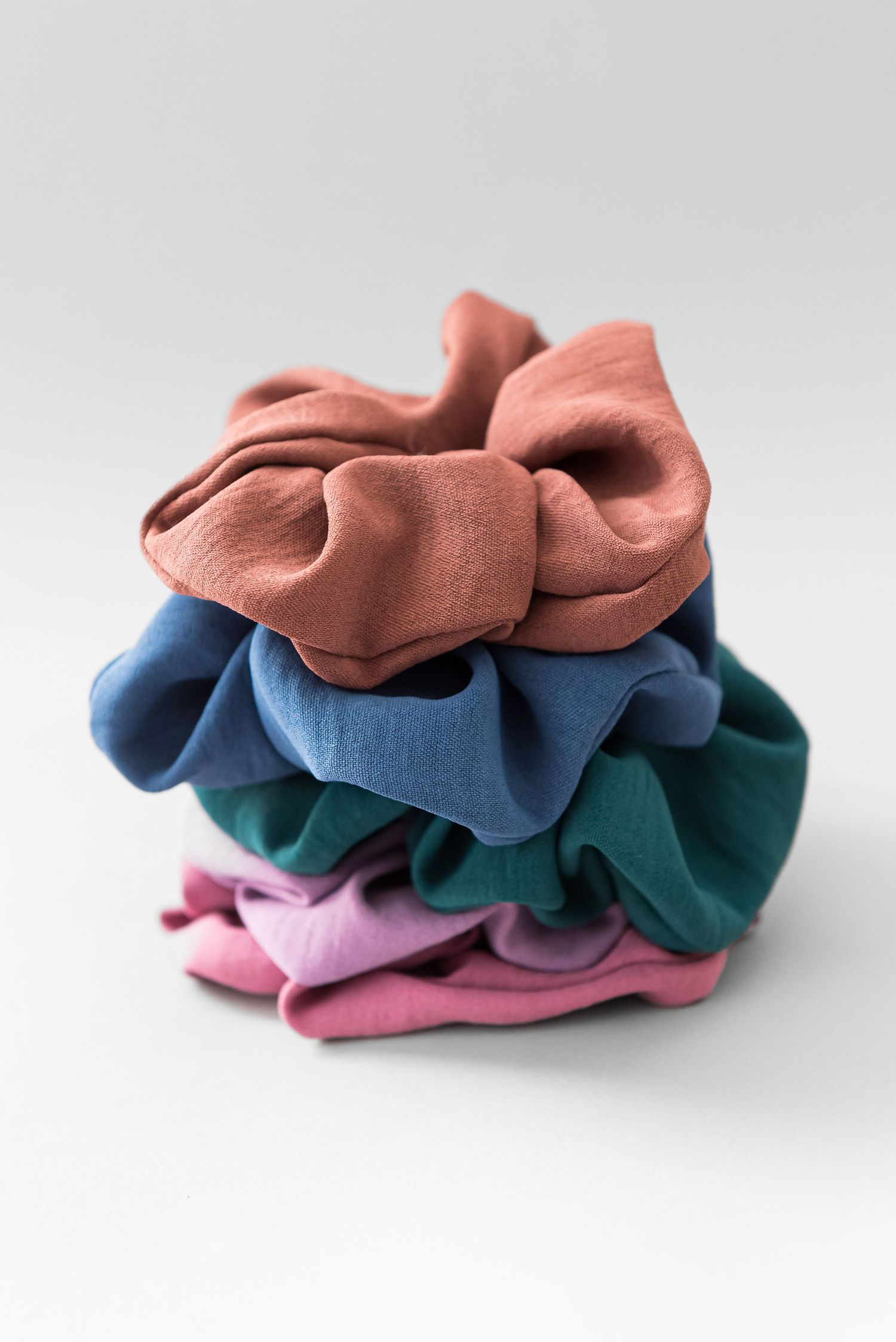 Stack of silk scrunchies in coral, navy, teal and pink colors on white background.