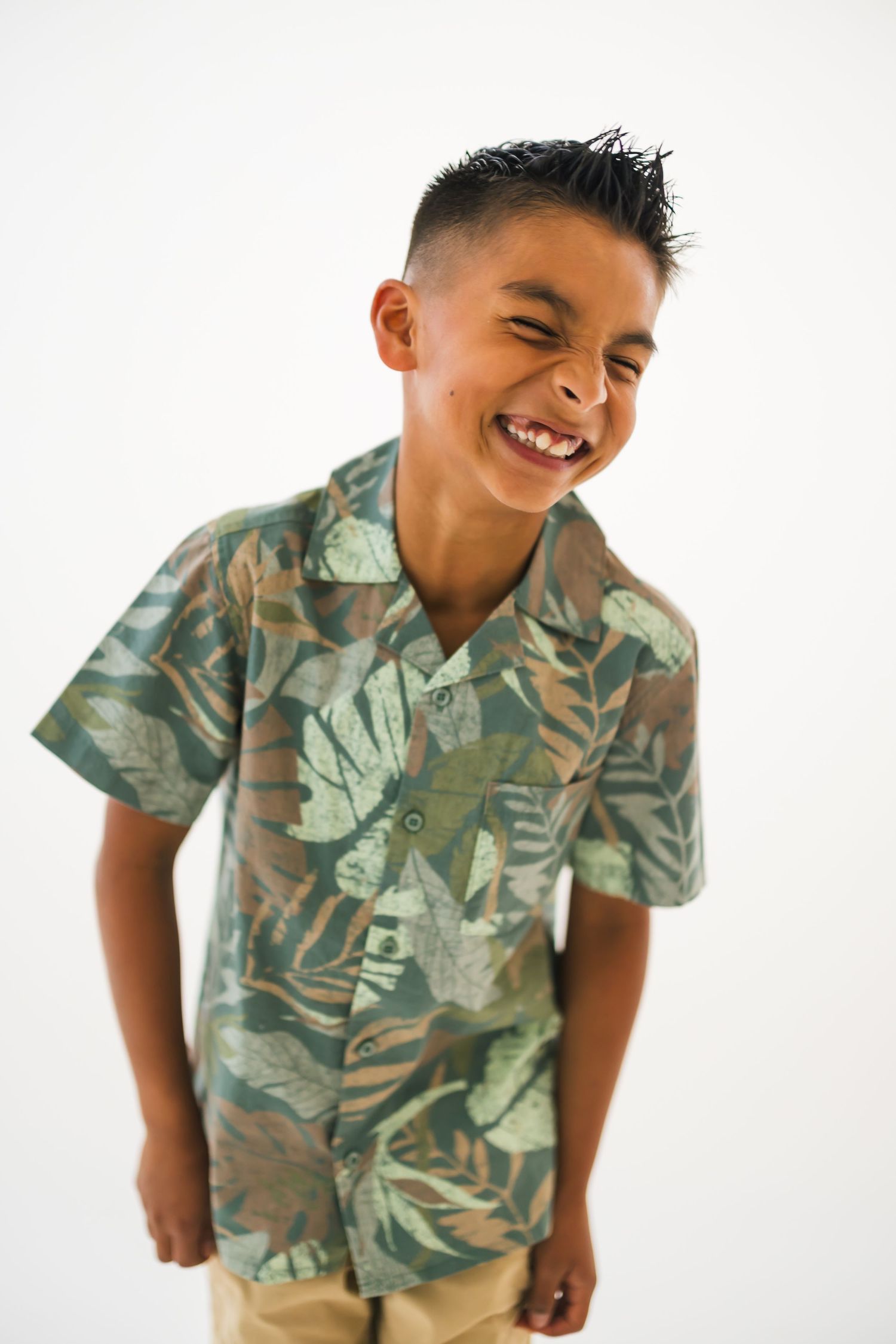 Young child wearing a tropical print Hawaiian shirt with leaf patterns smiles joyfully against white background.