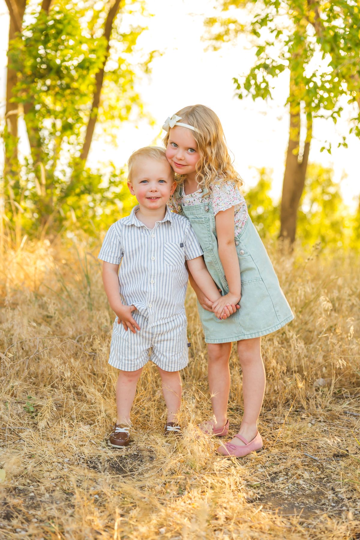 Children in summer outfits holding hands while standing in tall grass during golden hour.