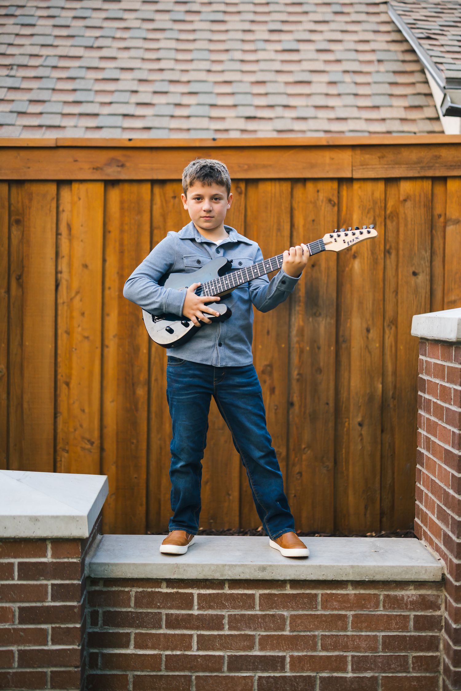 Young boy in denim plays electric guitar while standing on brick steps.
