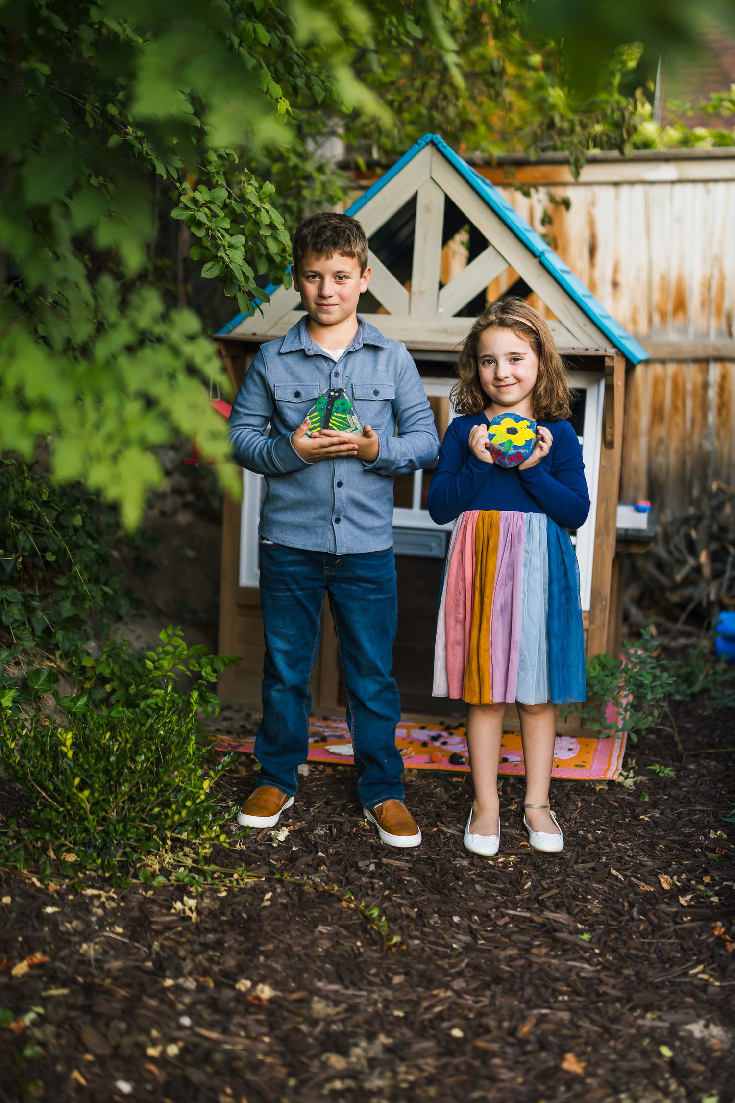Two children stand by a blue playhouse holding colorful toys in the garden.
