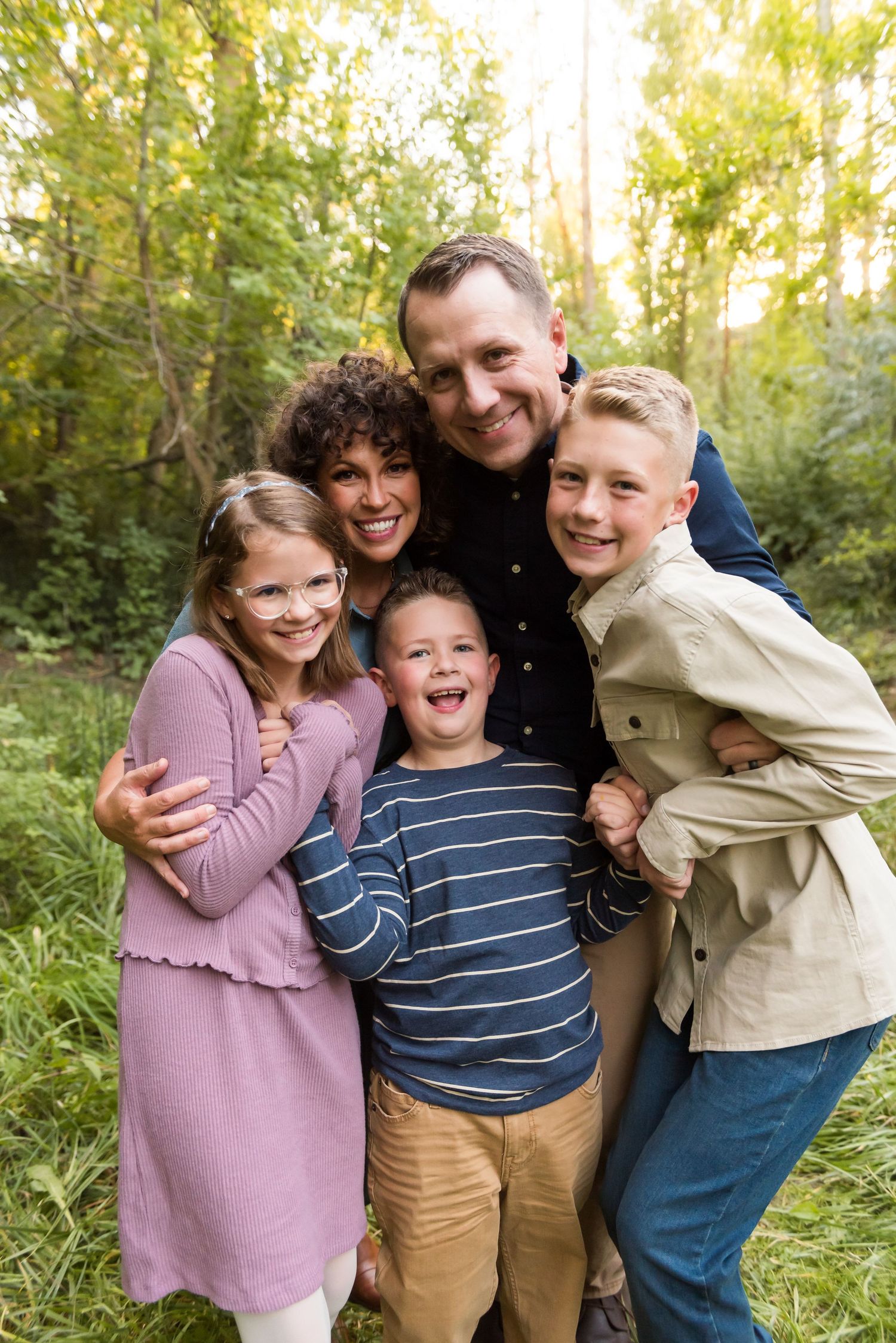 A family laughs while posing together outdoors in natural greenery during a photo session.