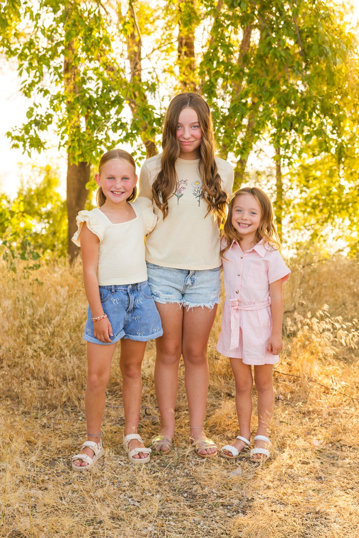 Three siblings stand together smiling in casual summer outfits during a golden hour photoshoot.