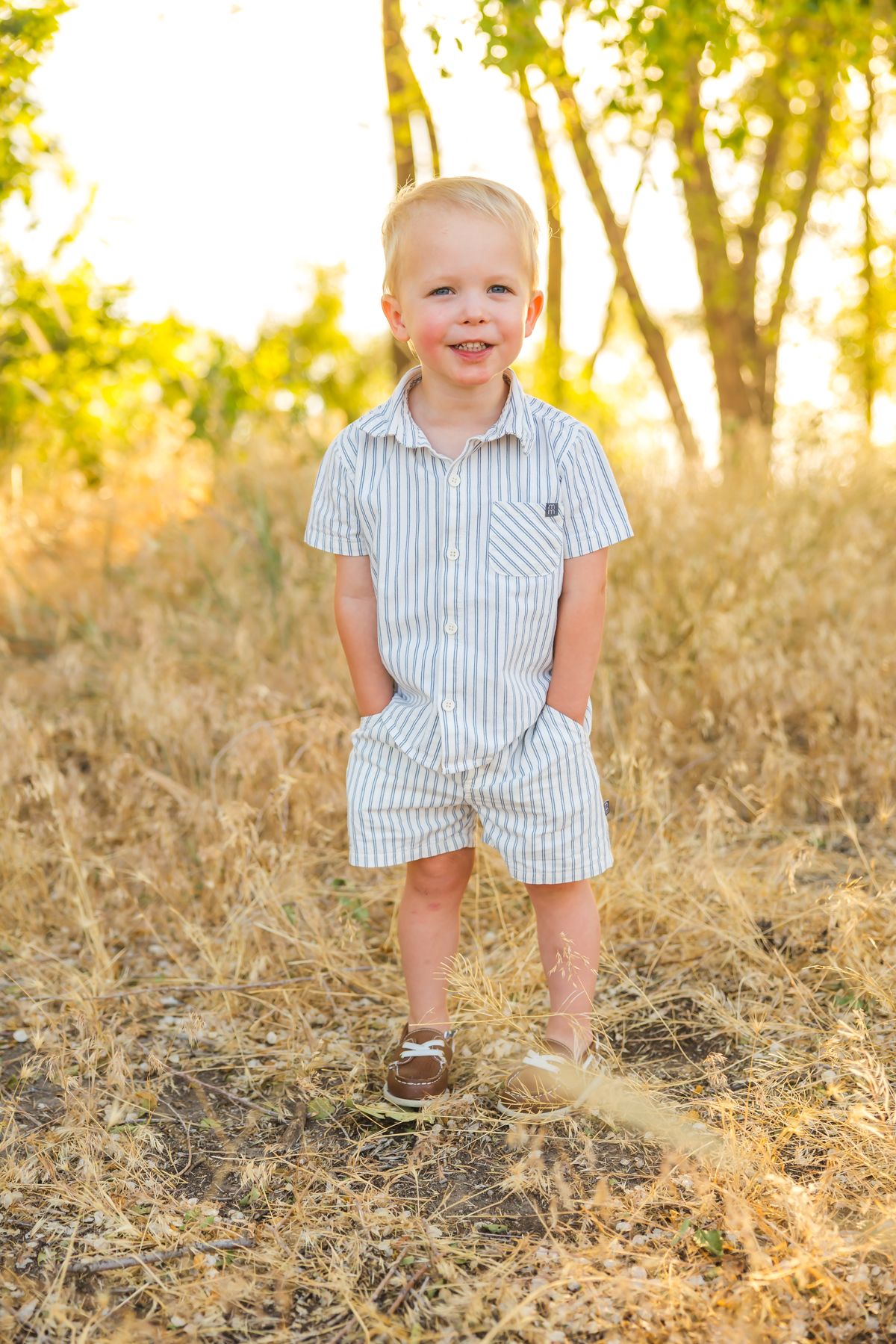 Child in striped outfit smiles while playing in grassy field during sunset.