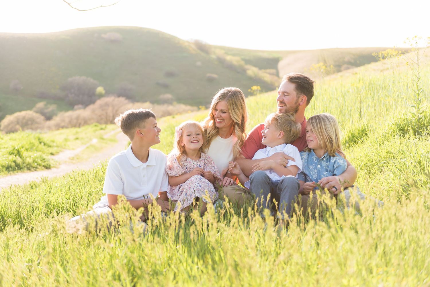 Family sitting together on sunny hillside meadow with rolling hills in background during golden hour.