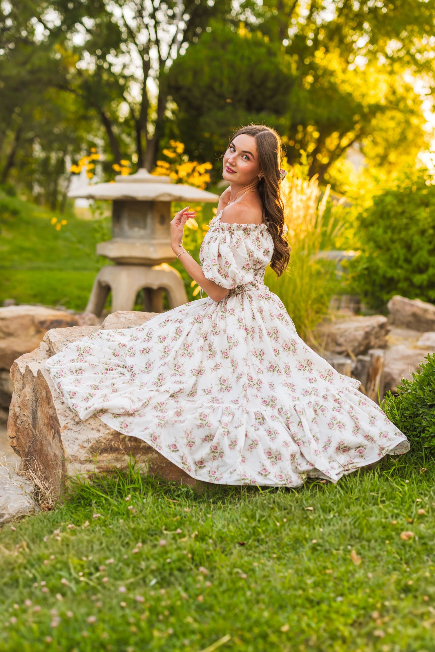 A floral white dress flows gracefully across green grass during a golden sunset photoshoot in a garden setting.