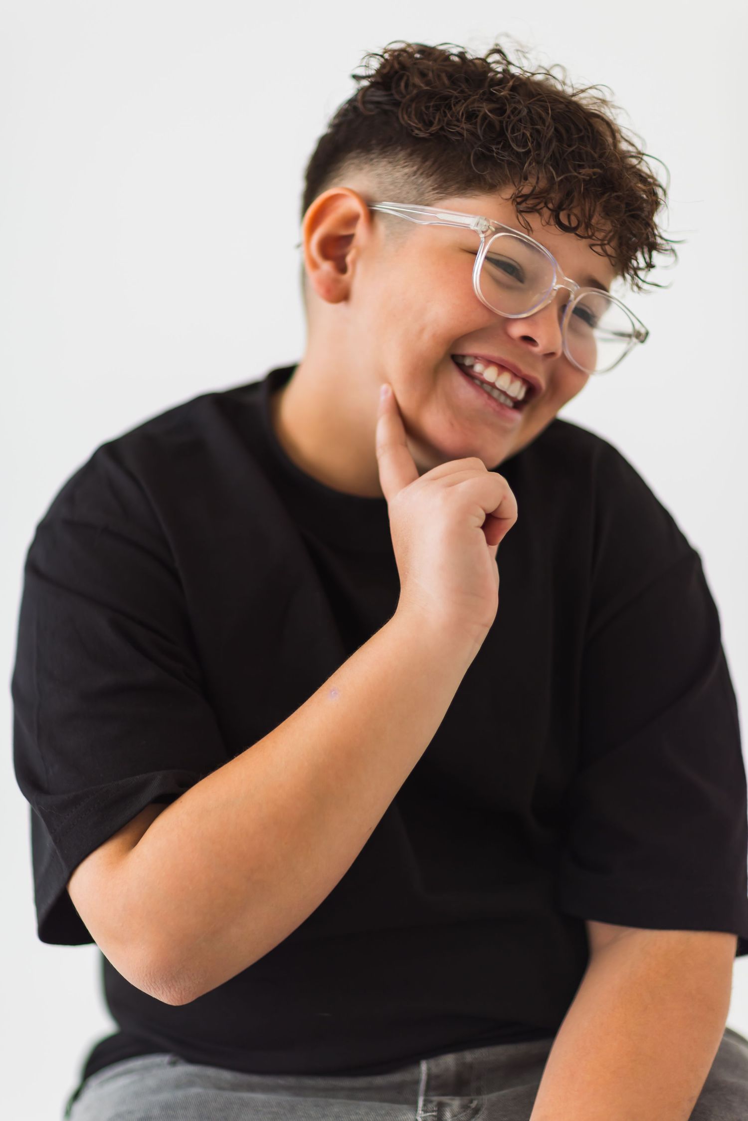 Person wearing clear glasses and black t-shirt smiles while resting chin on hand against white background.