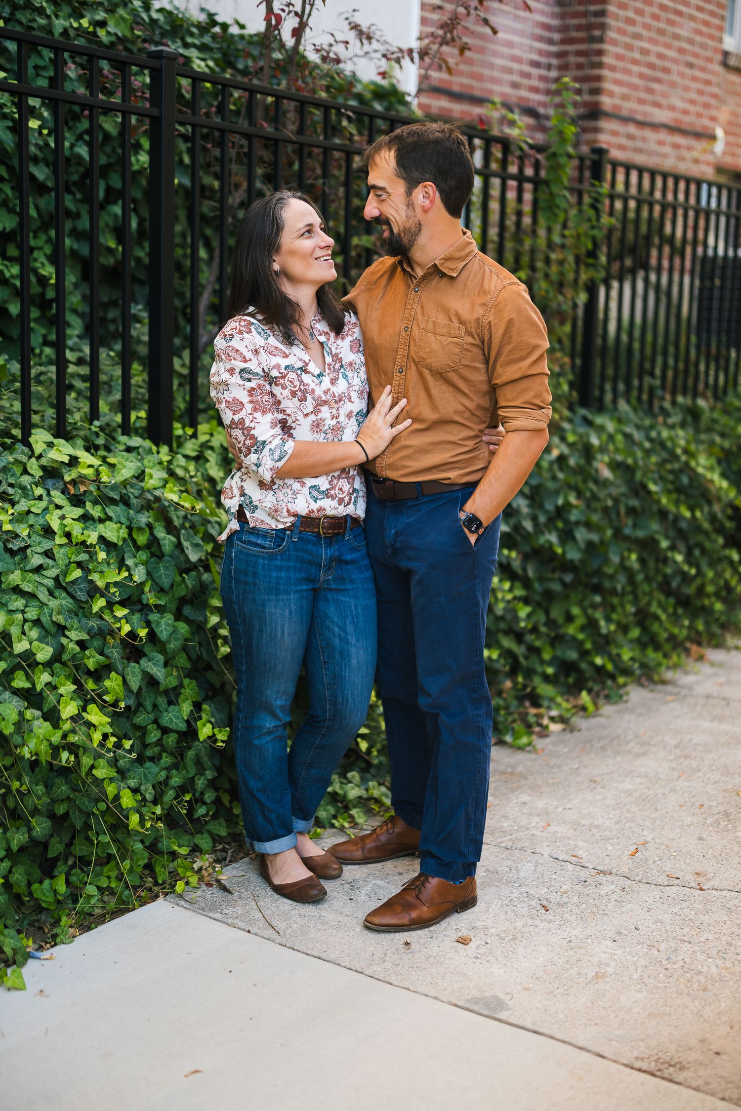 A couple shares a romantic moment against a brick wall and iron fence with lush greenery.