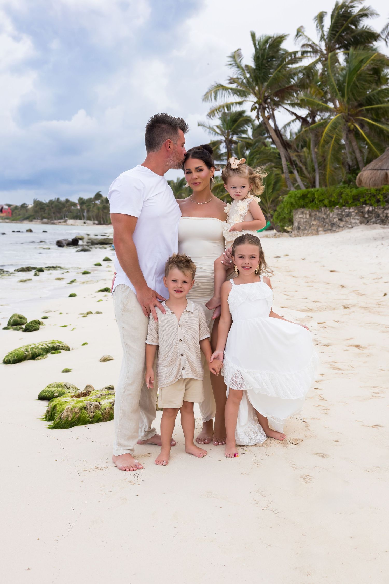 A family of five poses together on a tropical beach with palm trees and mossy rocks.