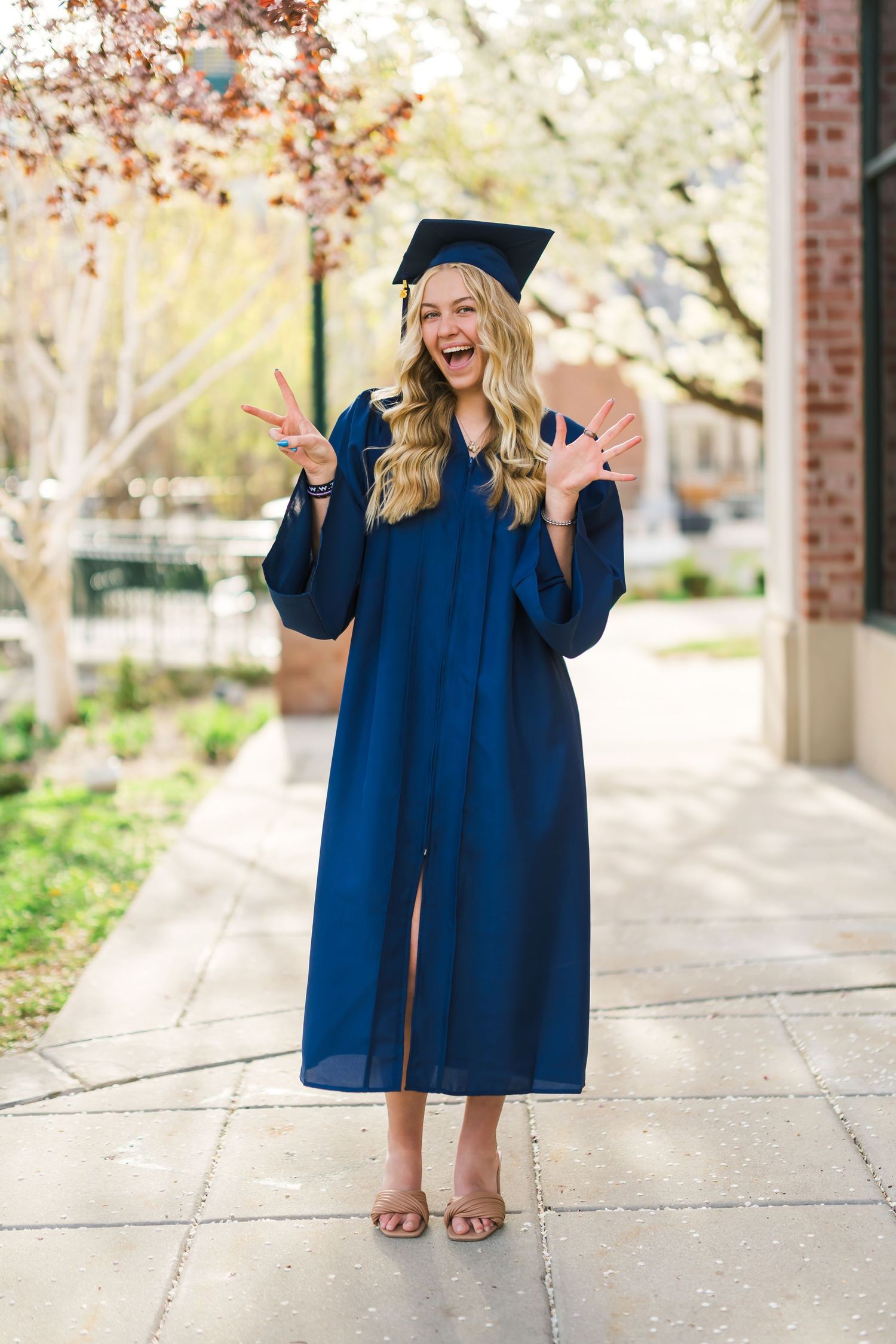 Graduate in blue cap and gown poses cheerfully on campus walkway with blooming trees. Senior portrait of Utah girl with spring blossoms