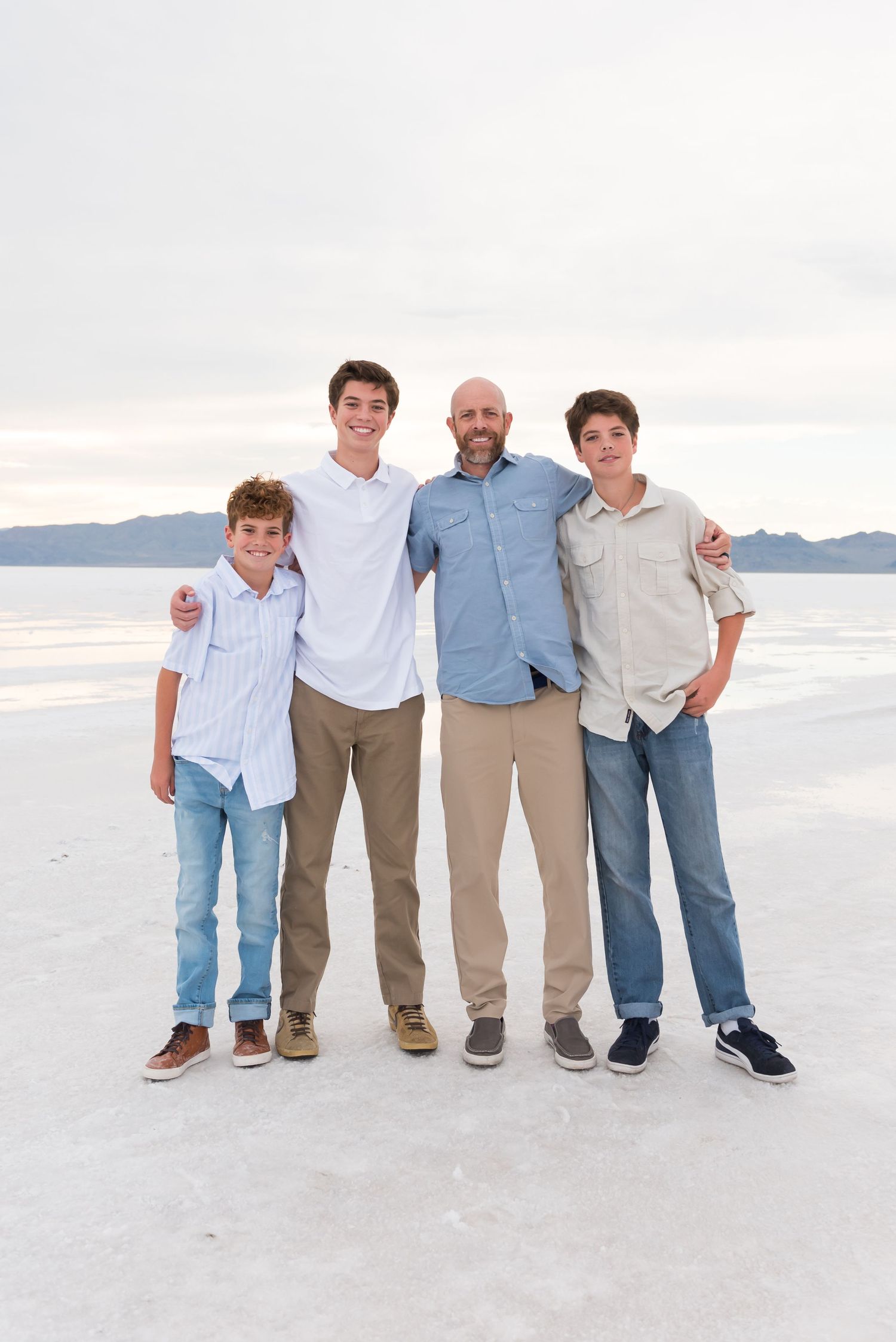 Family group wearing coordinated blue and white casual outfits posing together on white sand beach at sunset.