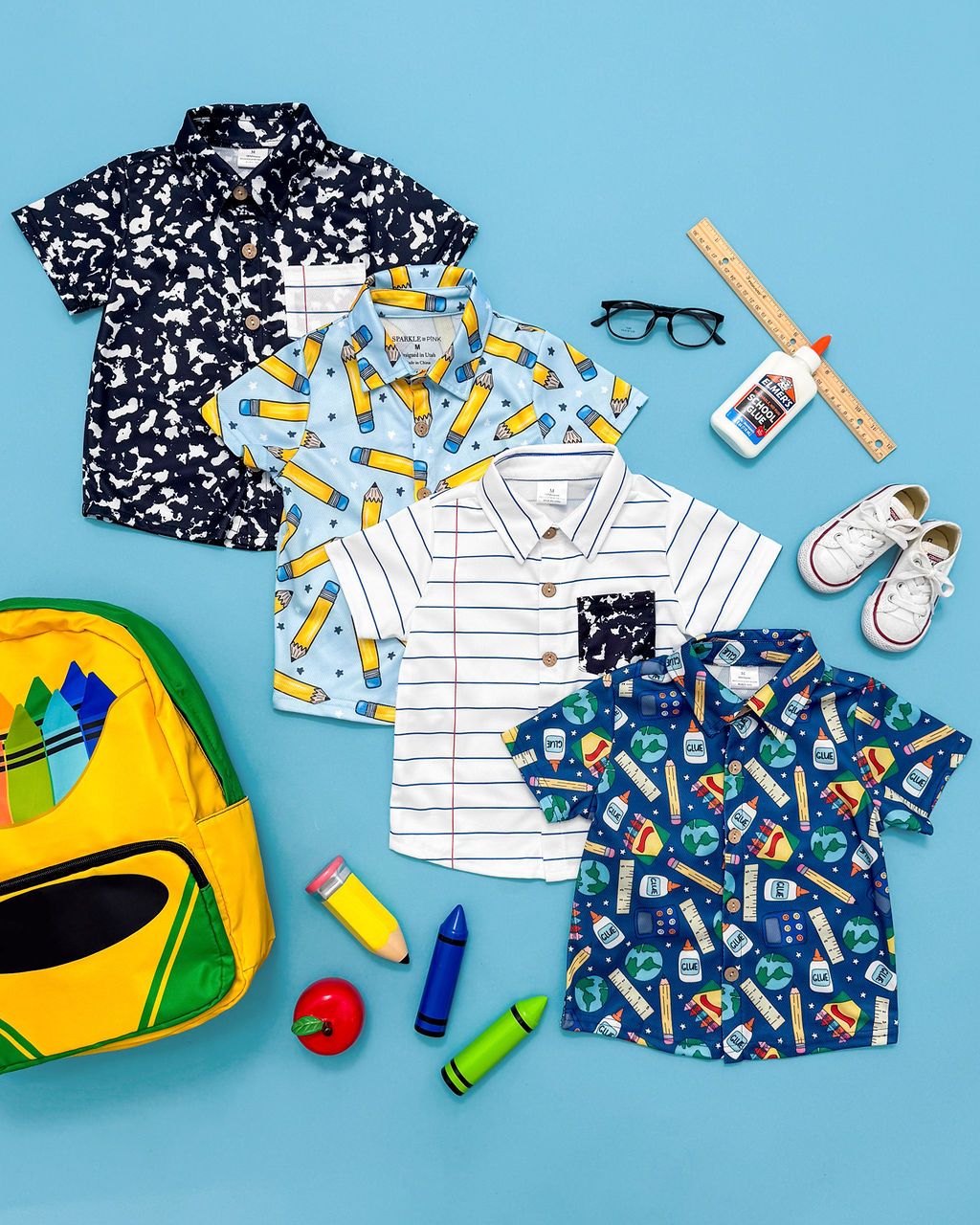 Children's clothing items including patterned shirts, school supplies, and accessories arranged on blue background in flat lay style.