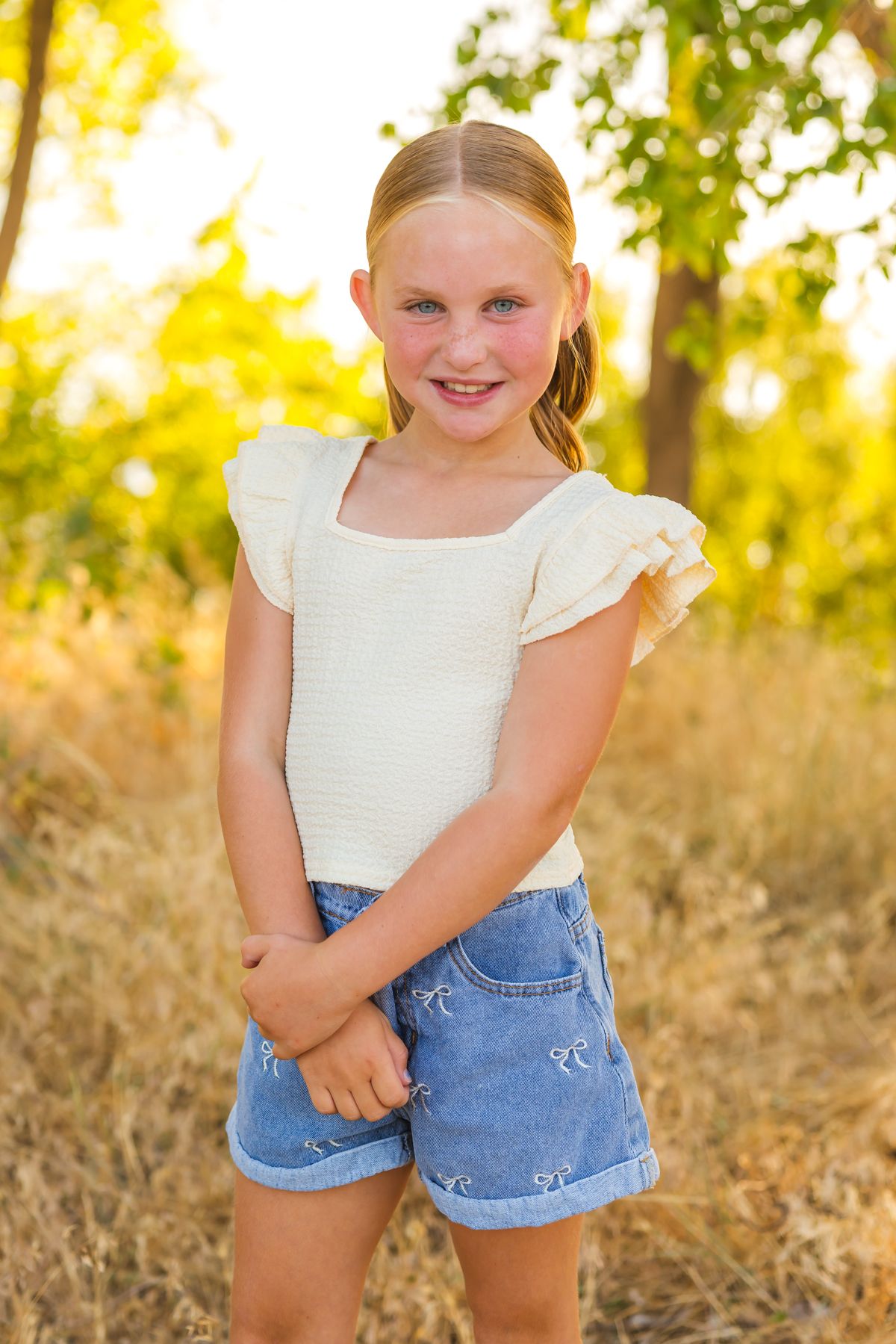 Portrait of a young person in a white ruffled top and denim shorts against a sunny nature backdrop.