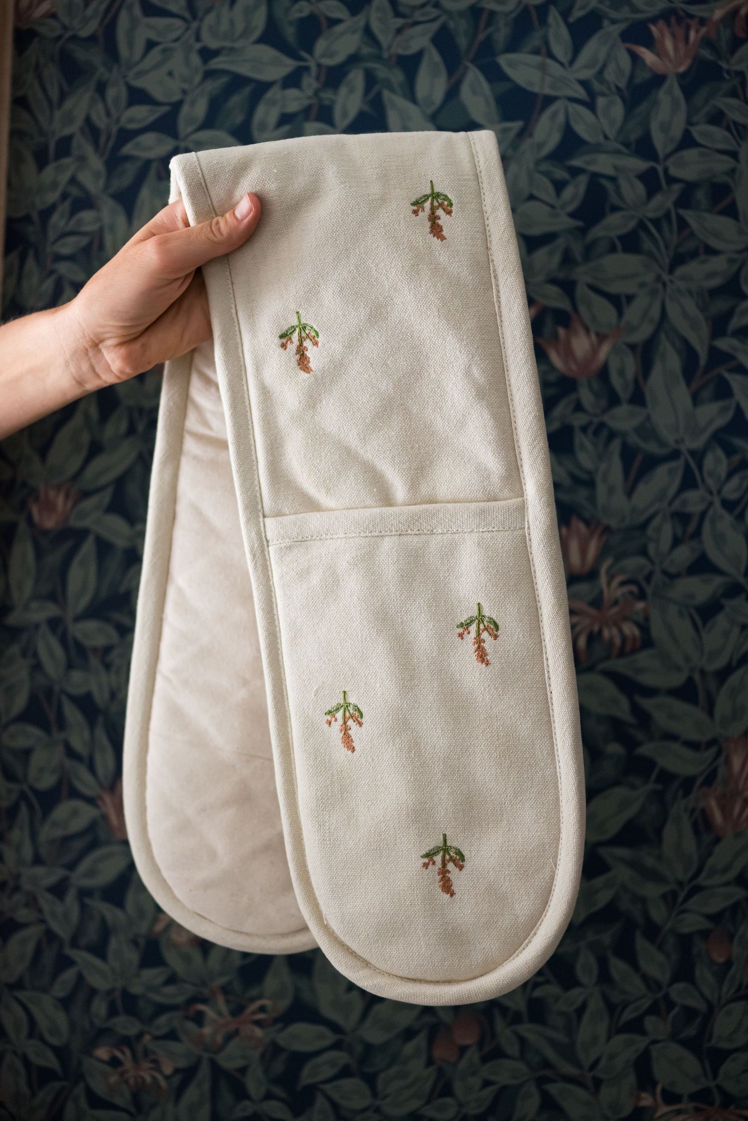 A white double oven mitt with embroidered palm tree designs being held against a dark floral background.