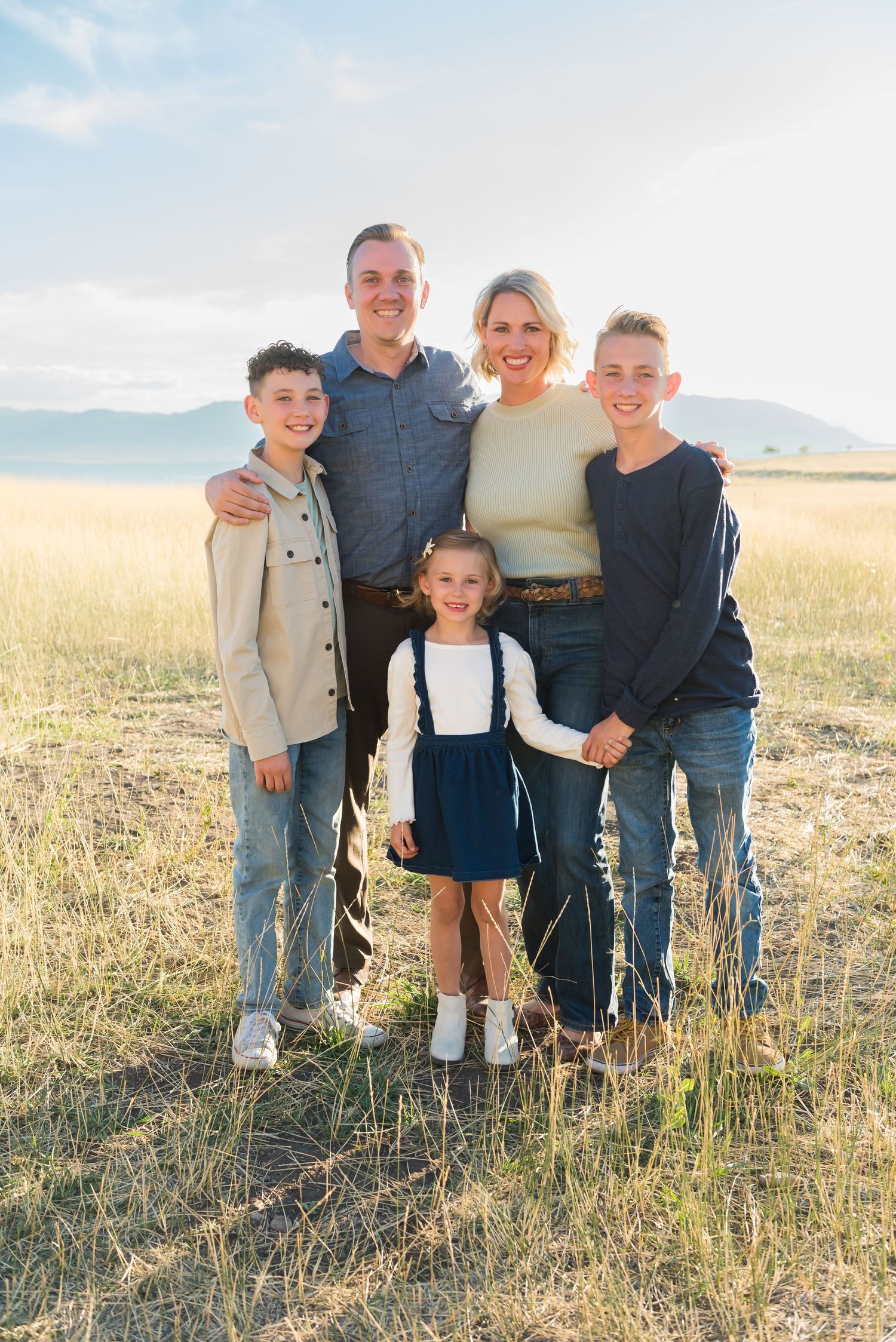 A family of five poses together in a golden field with mountains in the background.