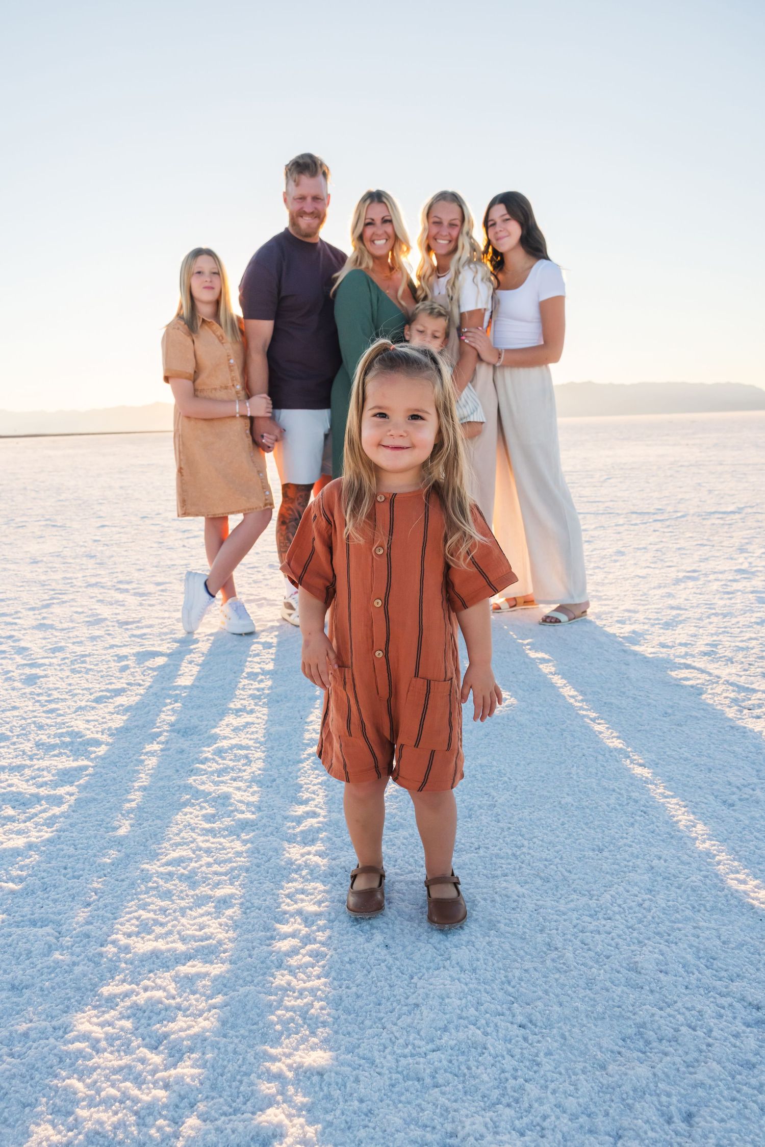 A smiling child in a rust-colored dress stands on white salt flats with family in the background.