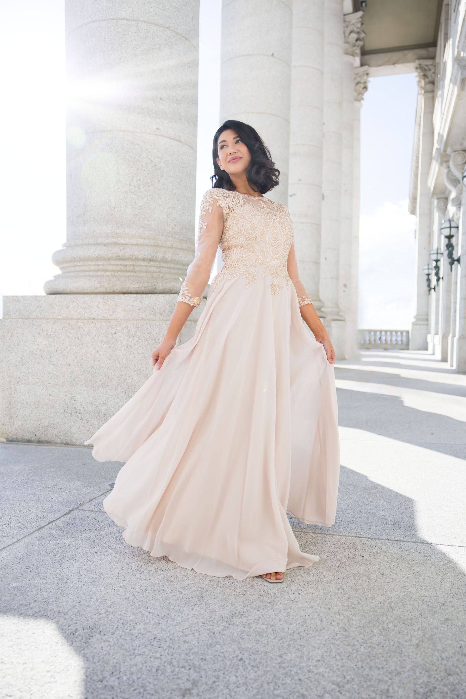 A flowing peach-colored formal dress is modeled in a bright architectural setting with classical columns.