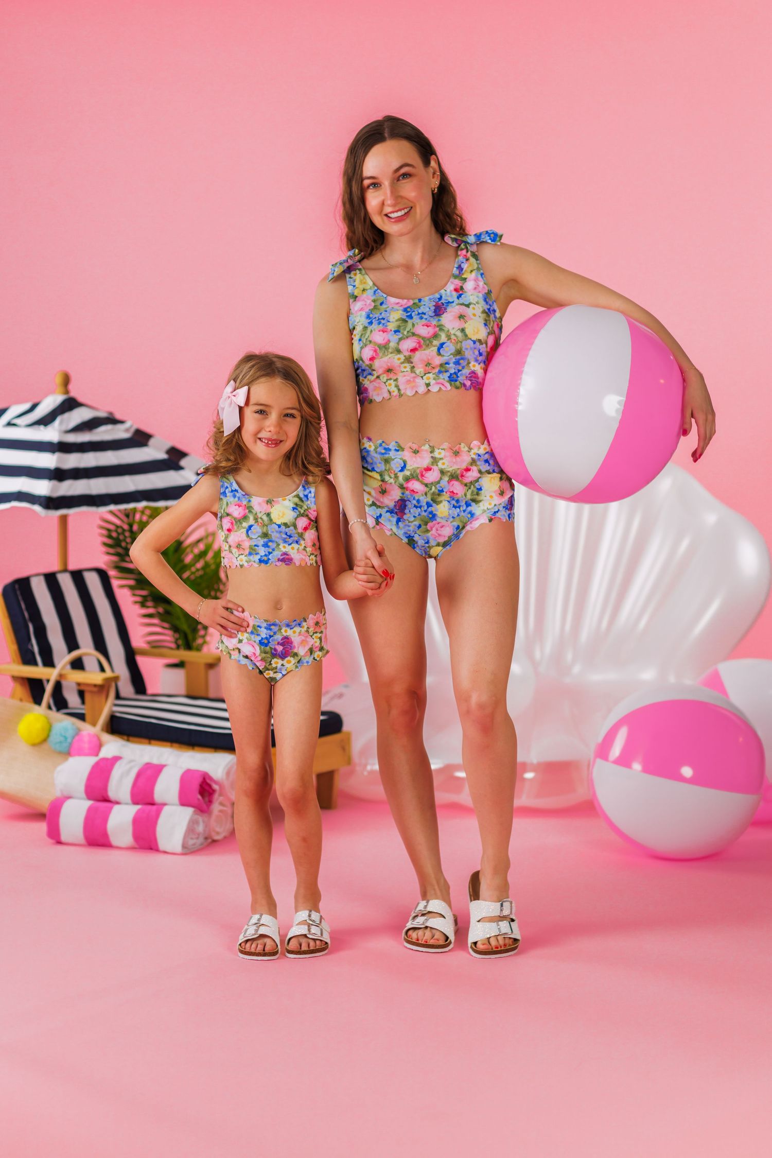 Matching floral swimsuits and pink beach accessories create a playful summer scene against a pink backdrop.