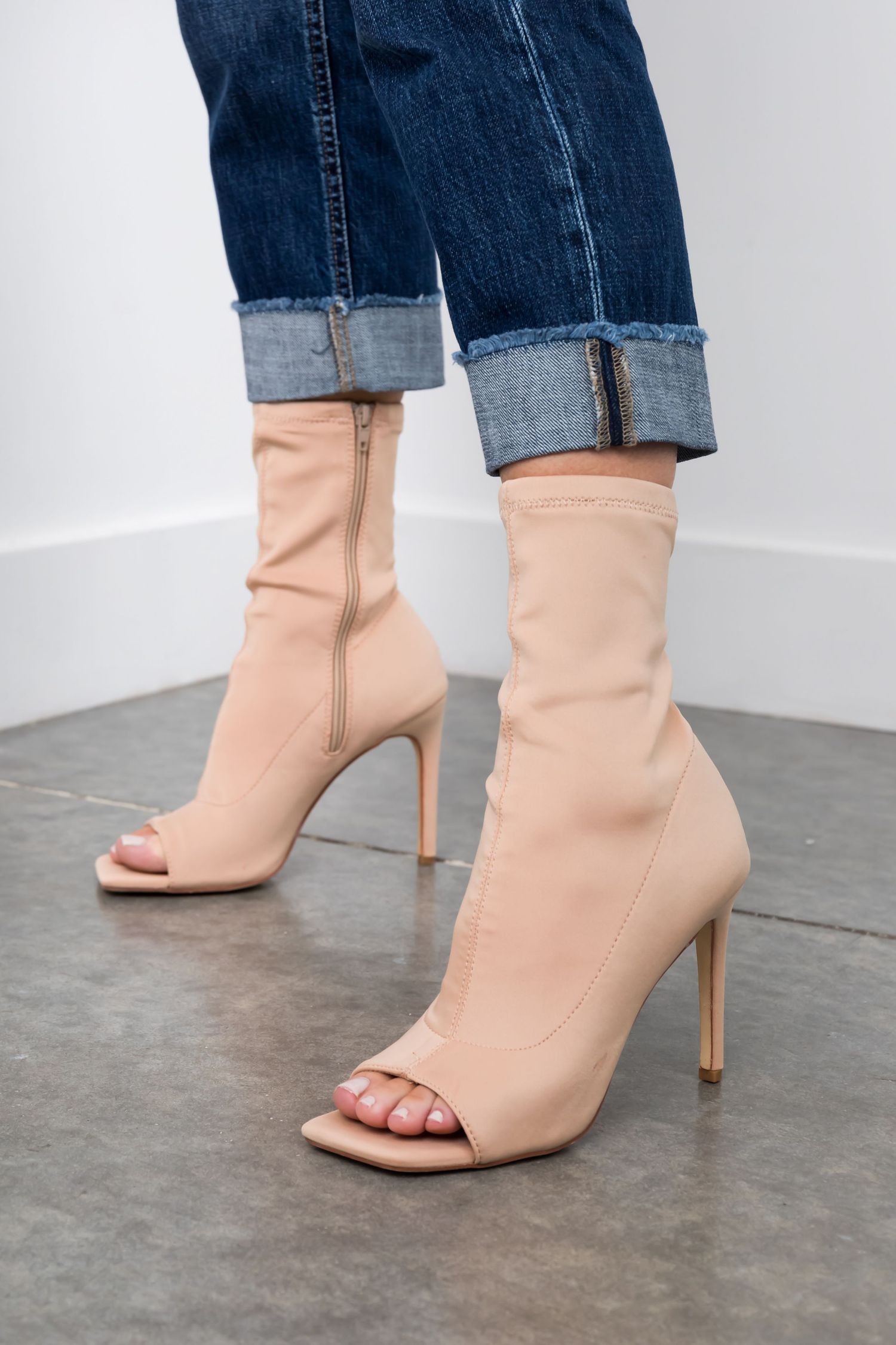 Person wearing rolled-up blue jeans and nude peep-toe ankle boots with stiletto heels stands on concrete floor.