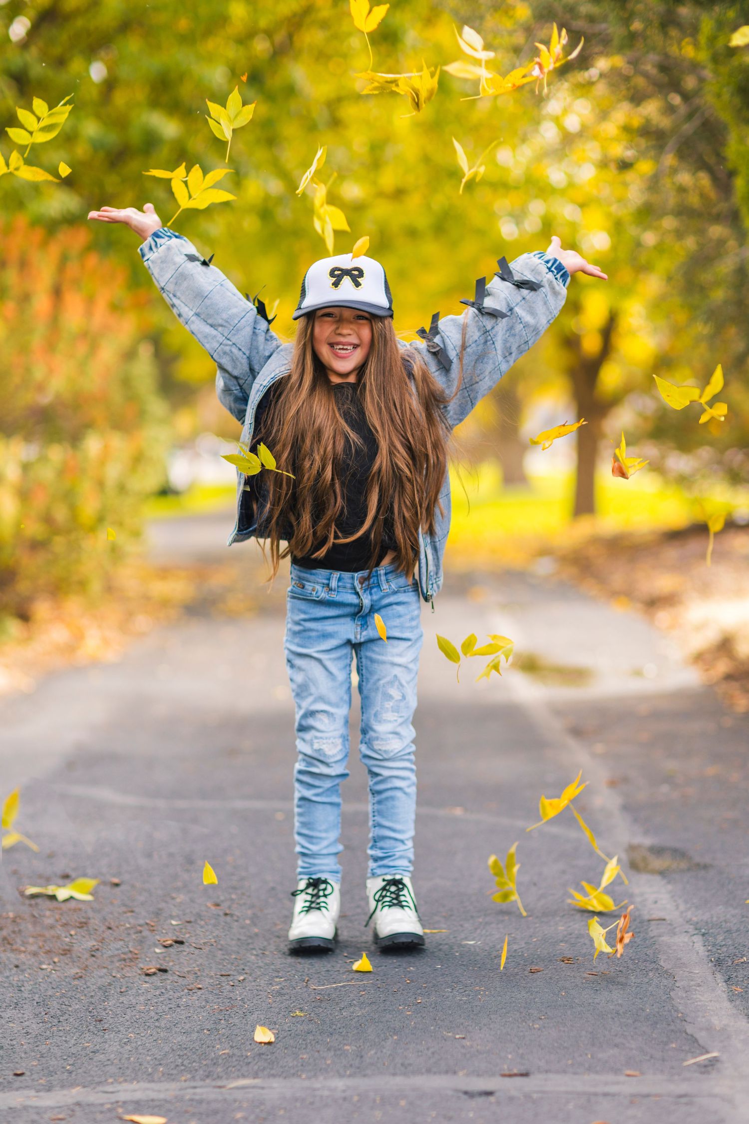A person in jeans and boots plays with falling autumn leaves on a path surrounded by golden foliage.
