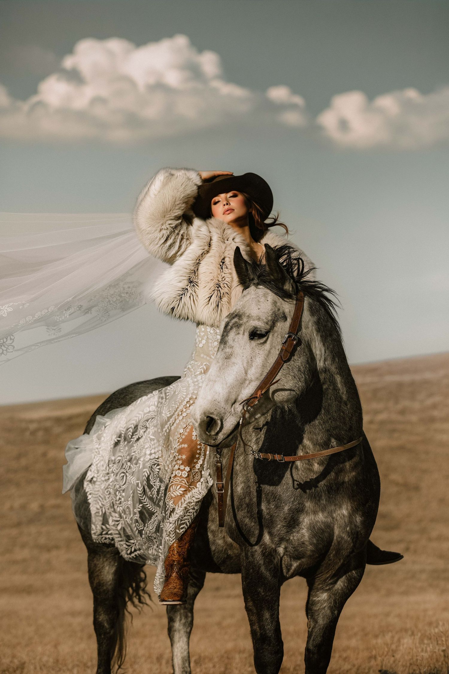 A woman in a lace bridal gown and cowboy hat rides a gray horse across an open field, her veil flowing in the wind.