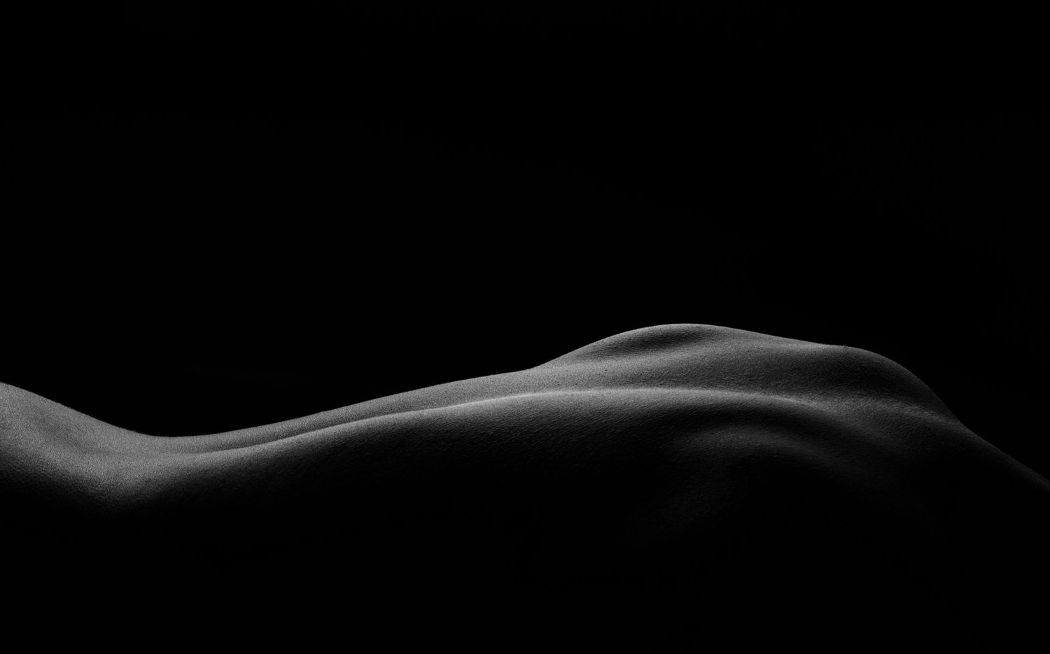 Black and white fine art nude photography highlighting the curves of the human body against a dark background.