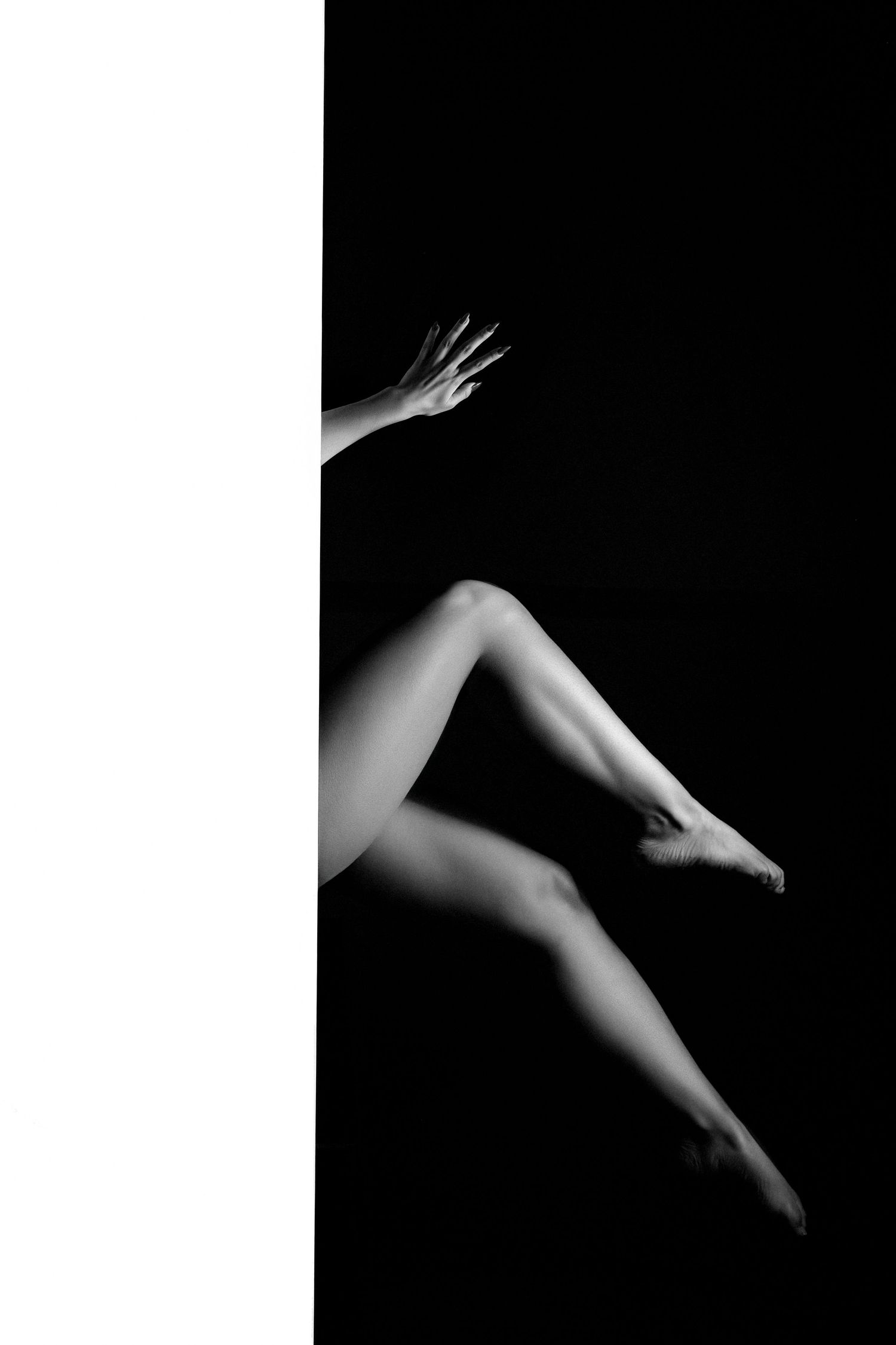 Black and white artistic photo of elegant female legs and hand against contrasting dark and light background.