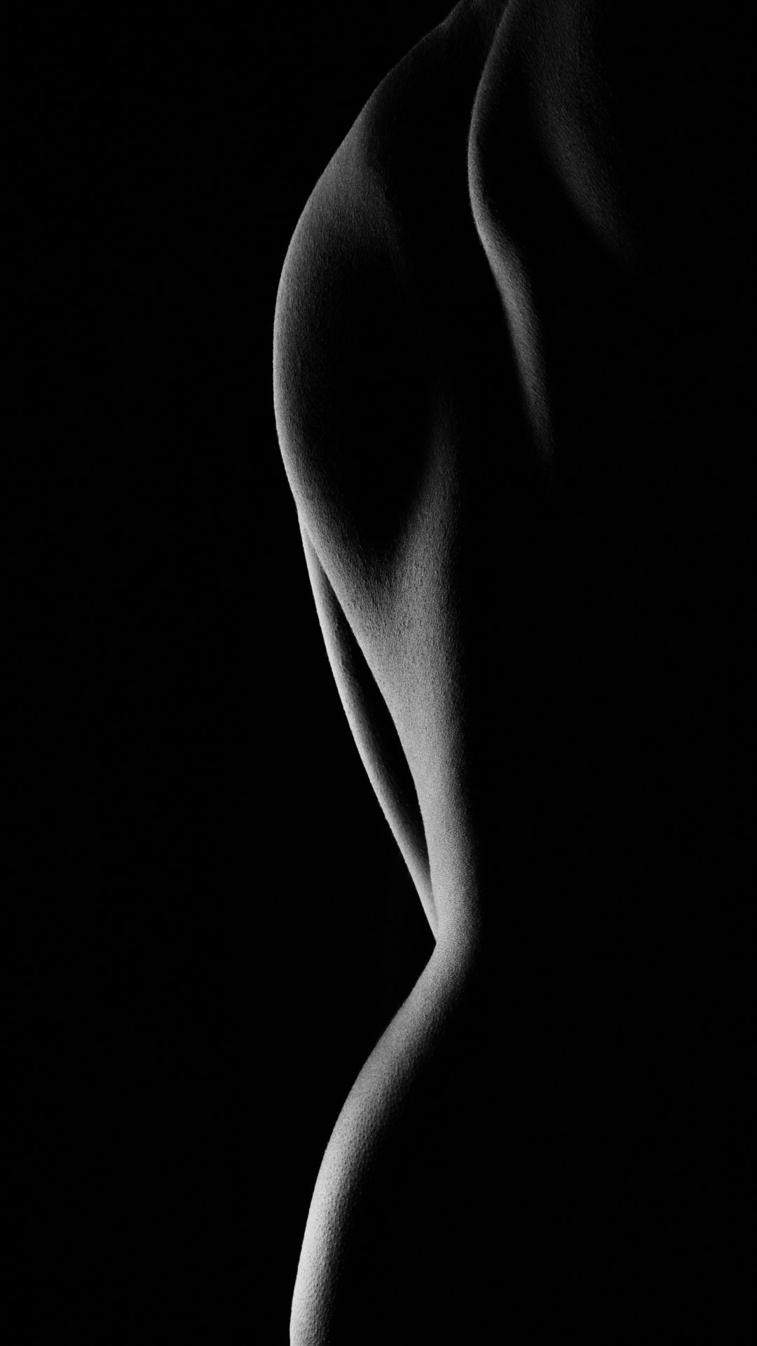 Black and white fine art nude photography showcasing elegant body curves with dramatic chiaroscuro lighting against dark background.