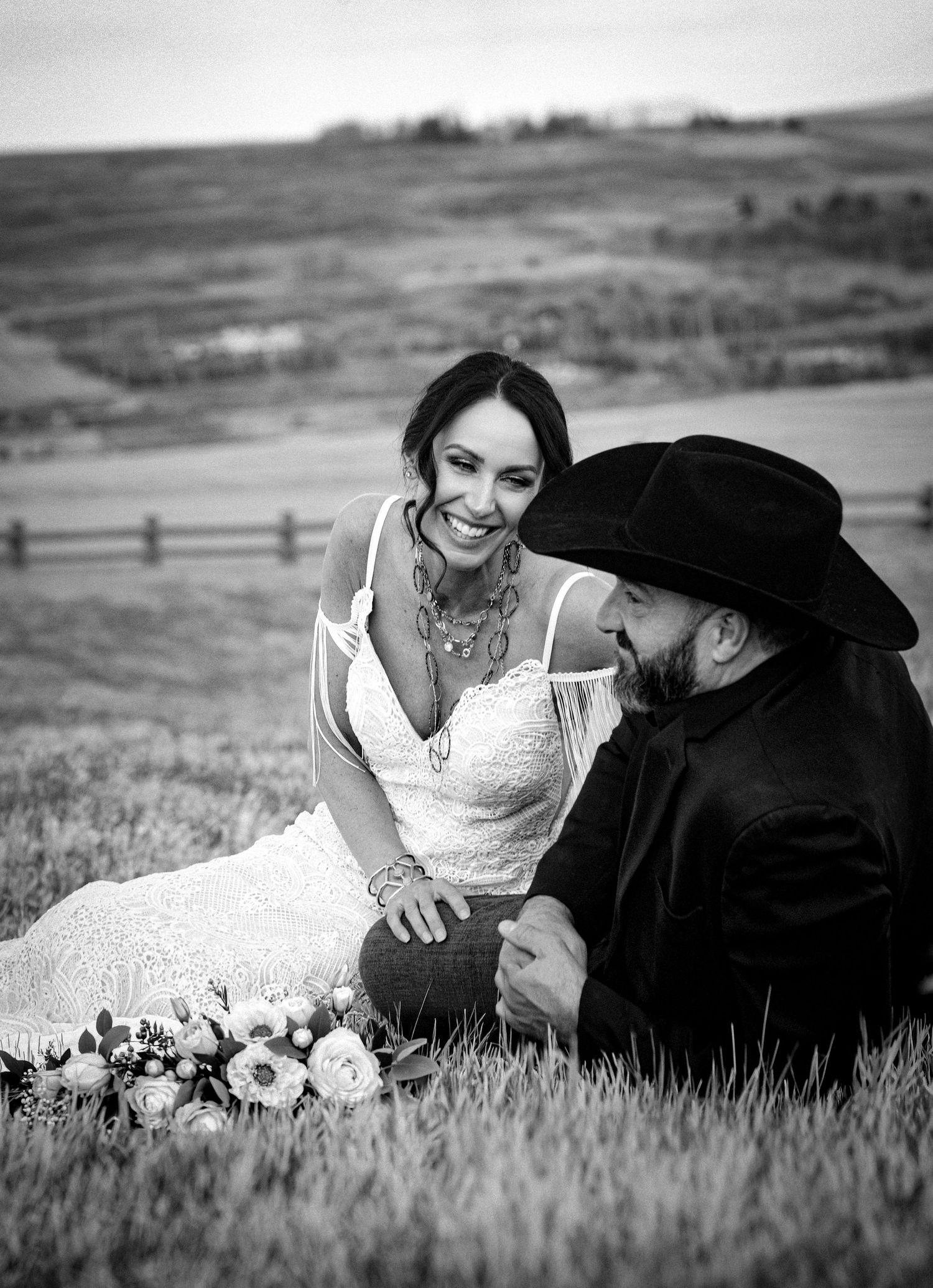Black and white photo of a smiling bride in lace dress sitting with groom in cowboy hat on a grassy hillside.