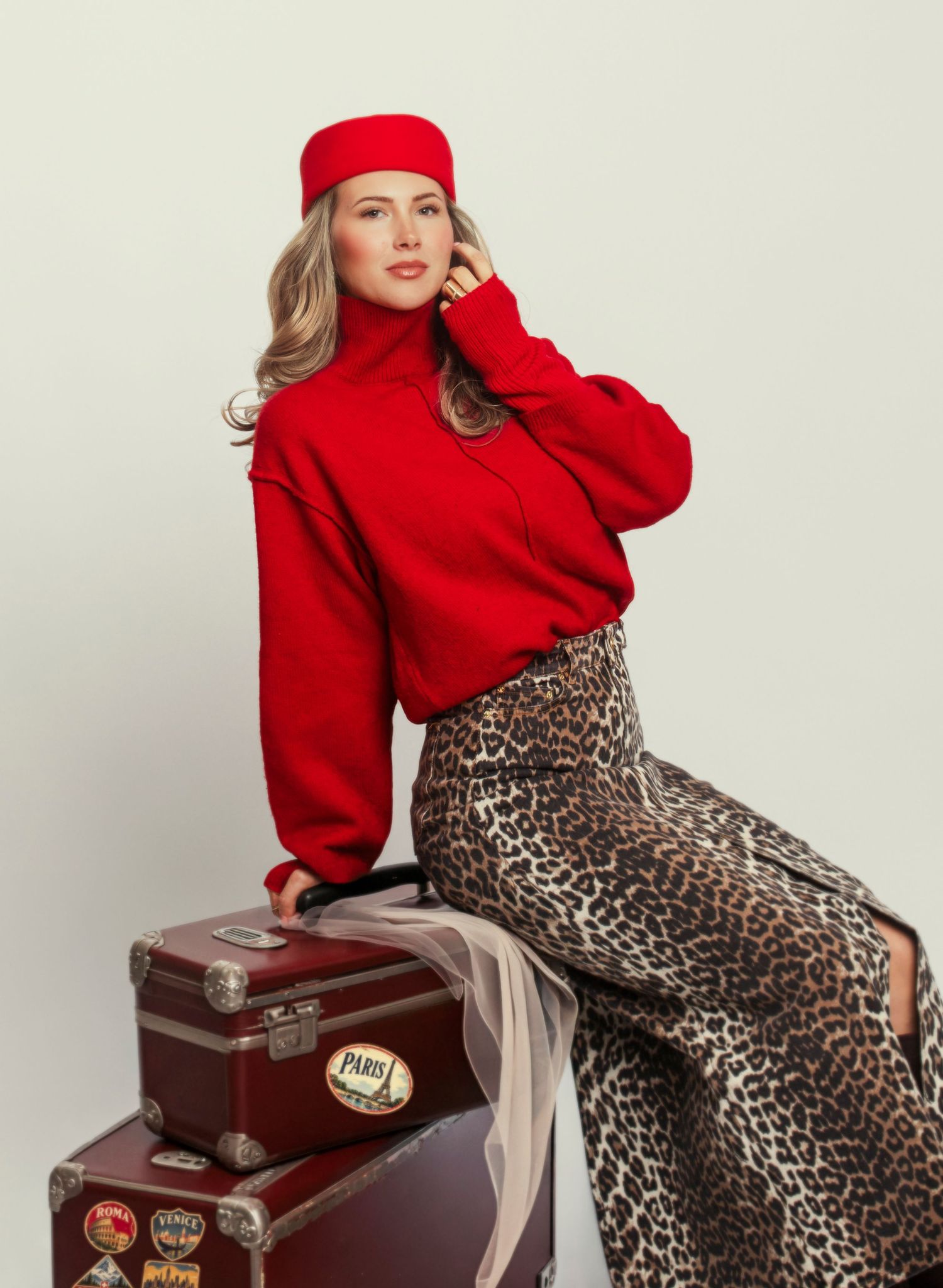 Woman in red turtleneck sweater and beret sits on vintage luggage wearing leopard print skirt.