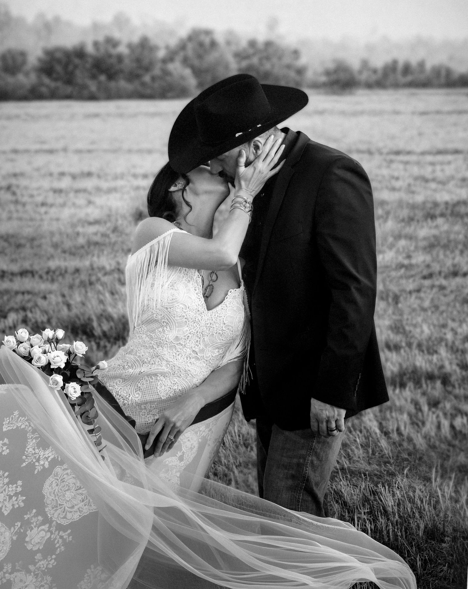 Black and white photo of a couple sharing a kiss in a field, bride in lace gown with veil, groom in cowboy hat.