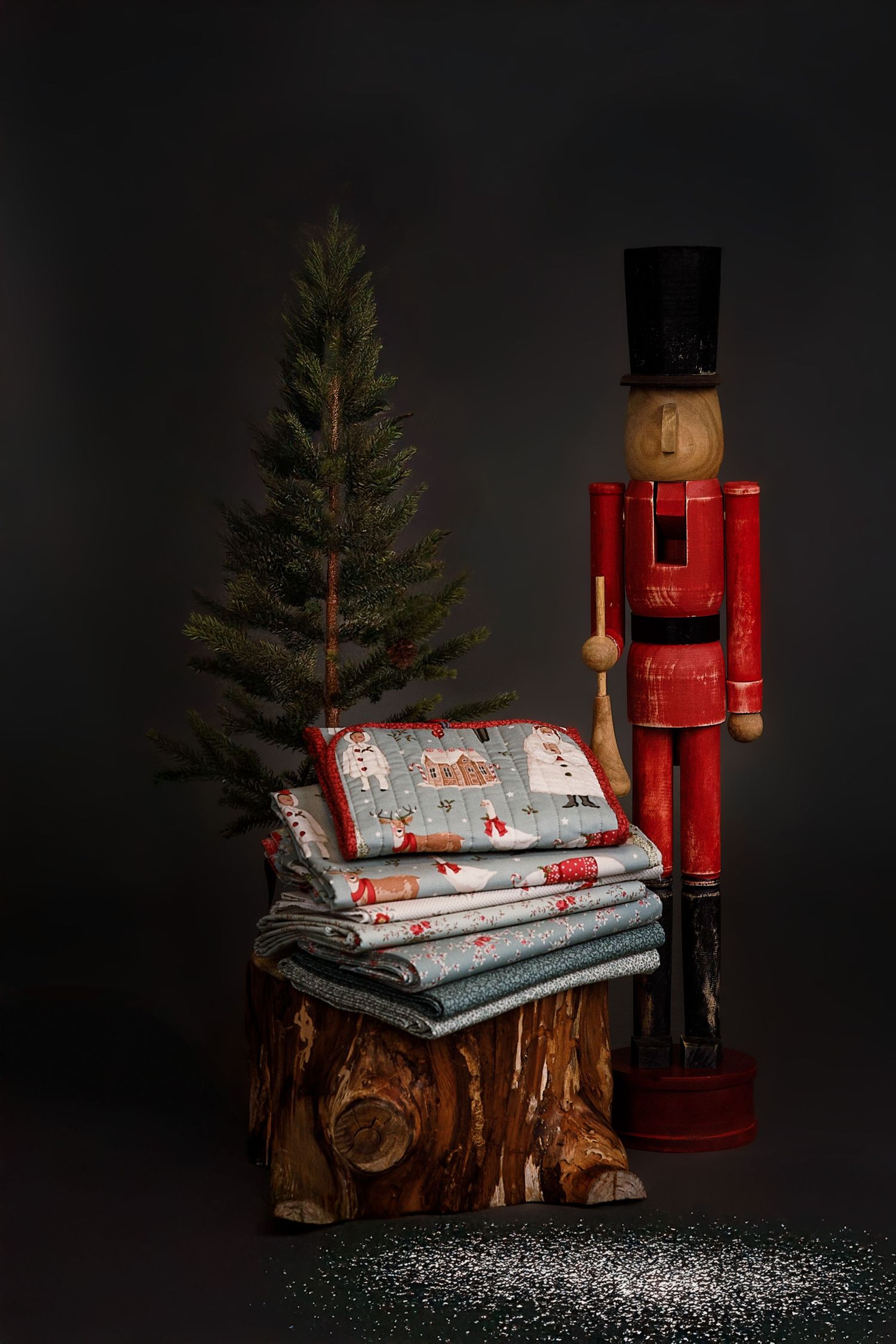 Christmas nutcracker figurine beside folded holiday quilts on a wood stump with a small pine tree on dark background.