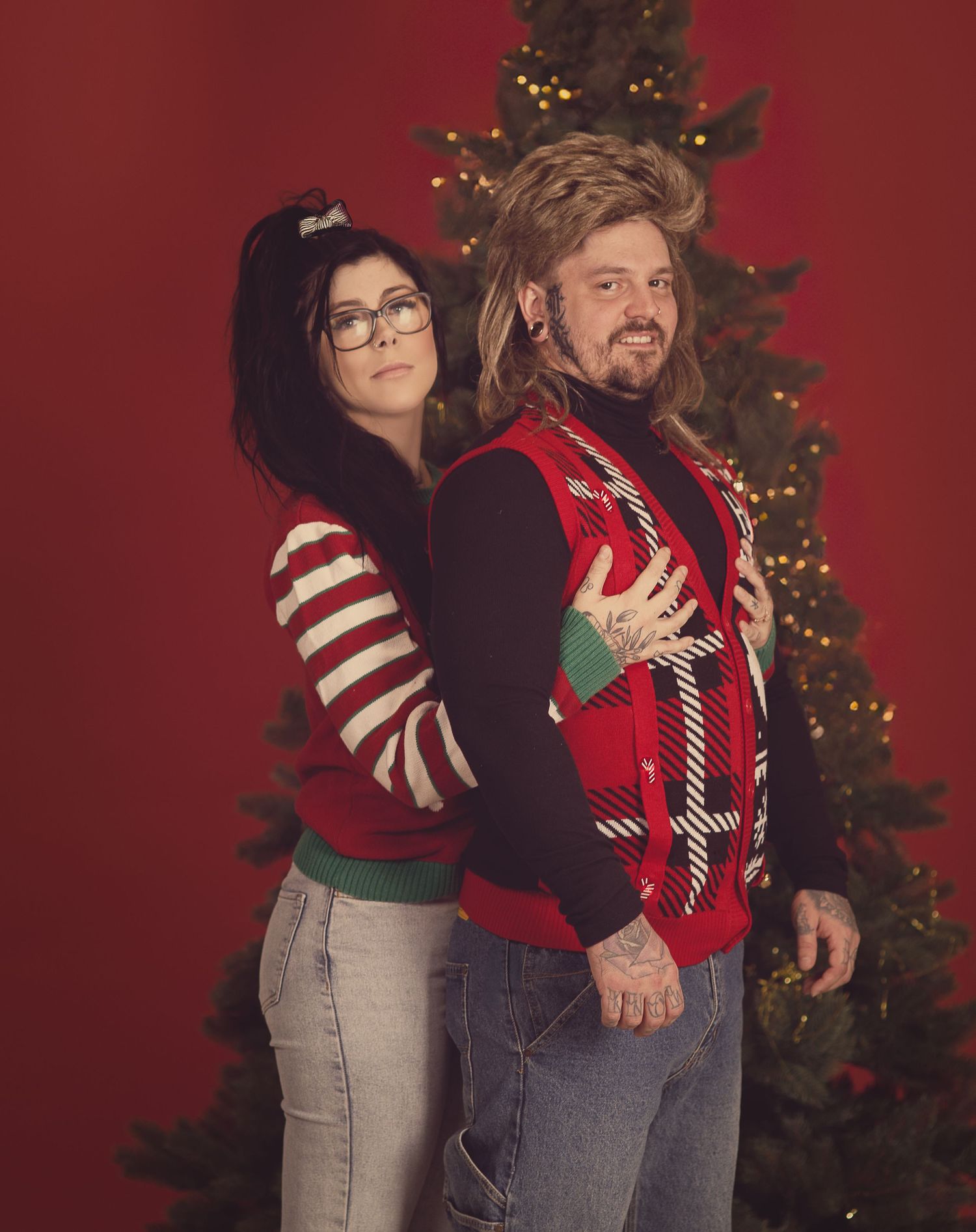Couple in a retro Christmas photo wearing festive sweaters, posing in front of a decorated tree against a red backdrop.