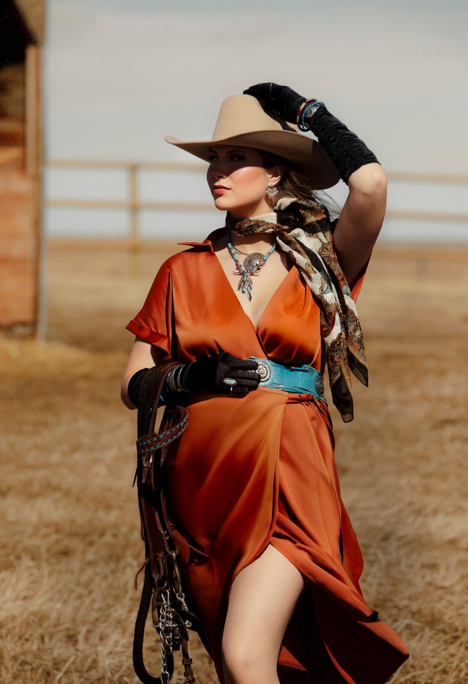 Western-inspired fashion photo with orange wrap dress, cowboy hat, turquoise belt and black gloves in desert setting.